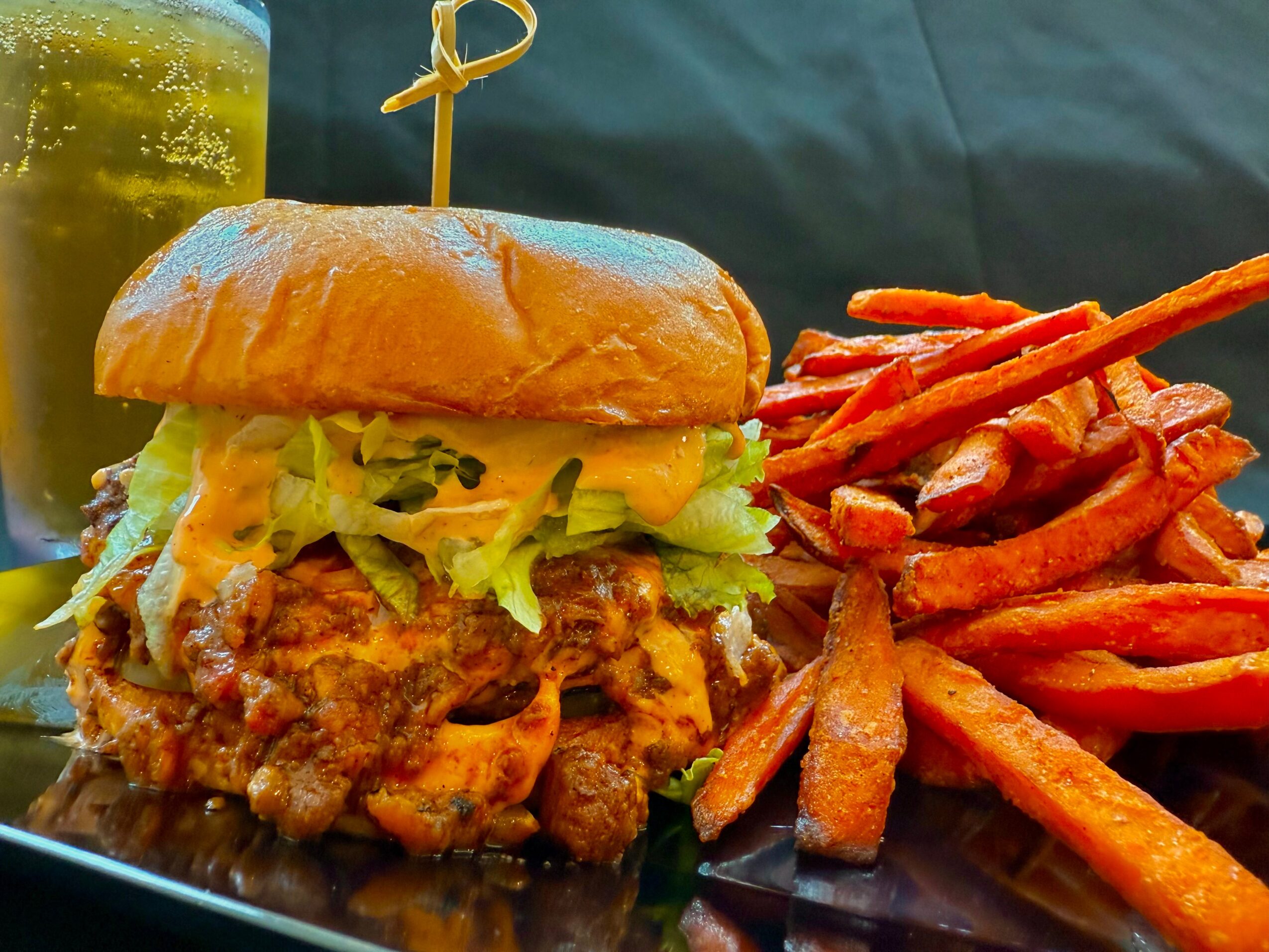 Burger with lettuce, cheese, and fried chicken, served with sweet potato fries on a black plate.