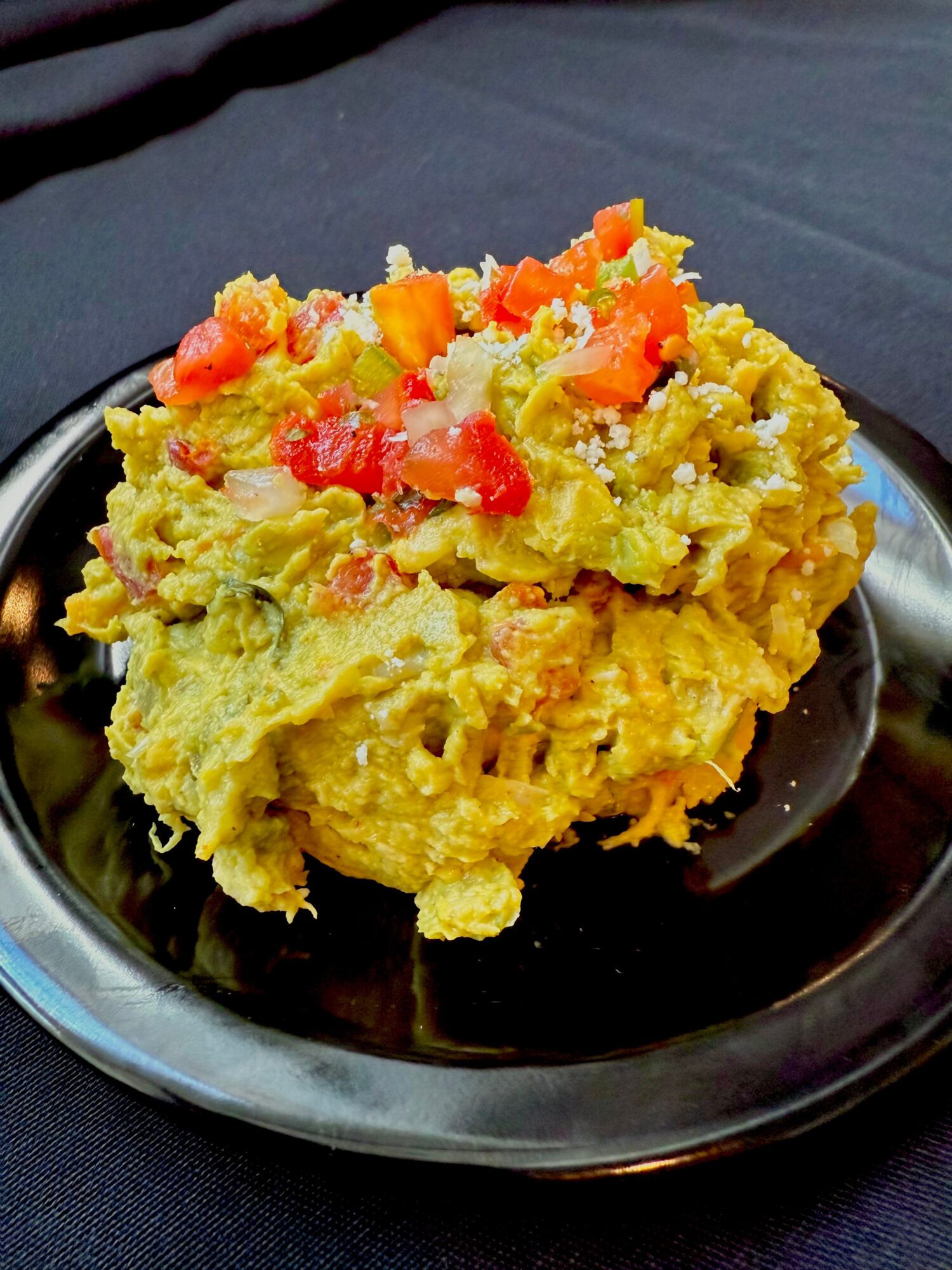 A serving of guacamole topped with diced tomatoes on a black plate, placed on a dark surface.