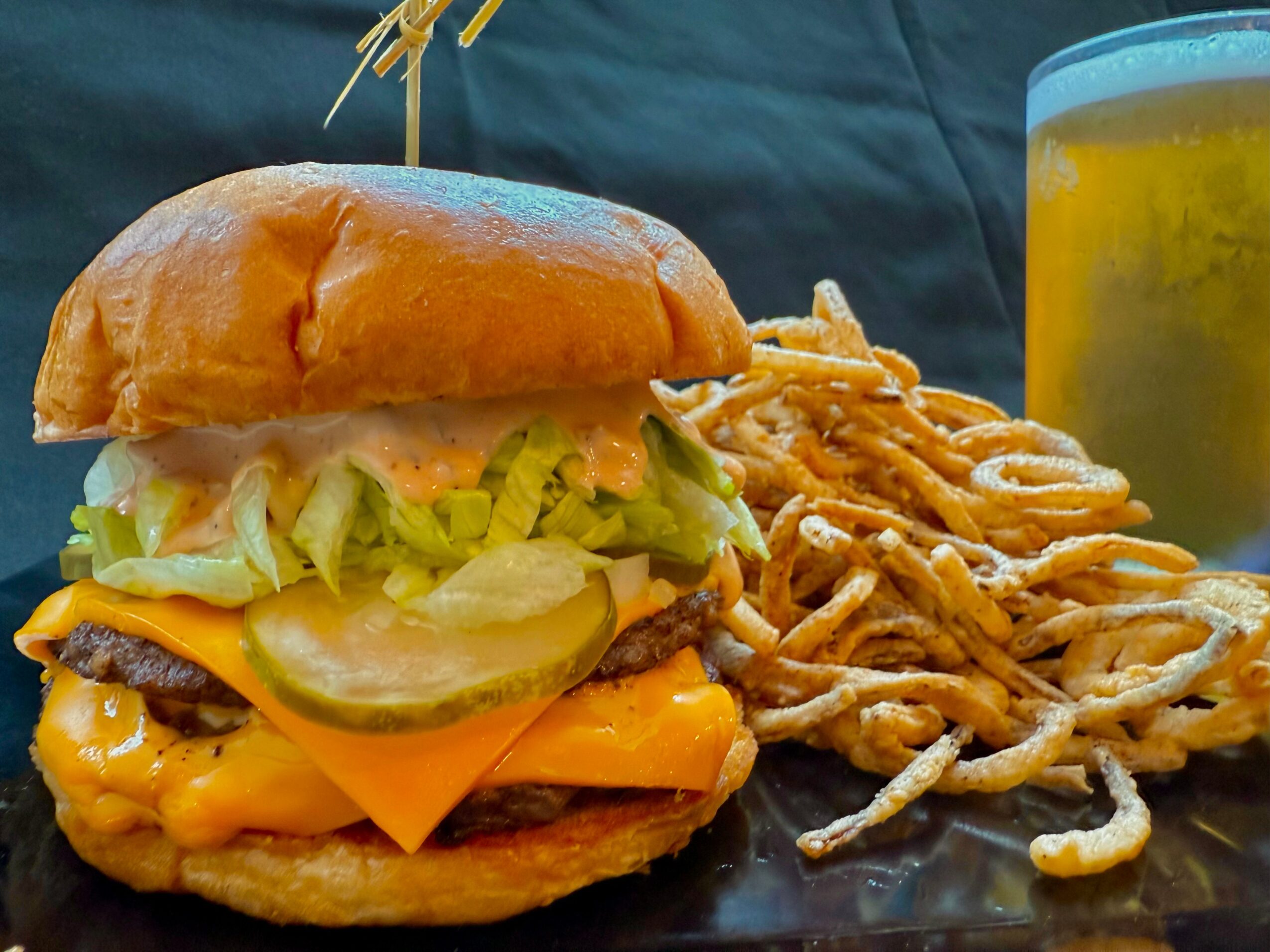 A burger with lettuce, pickles, cheese, and a bun, served with fried onion strings and a glass of beer.