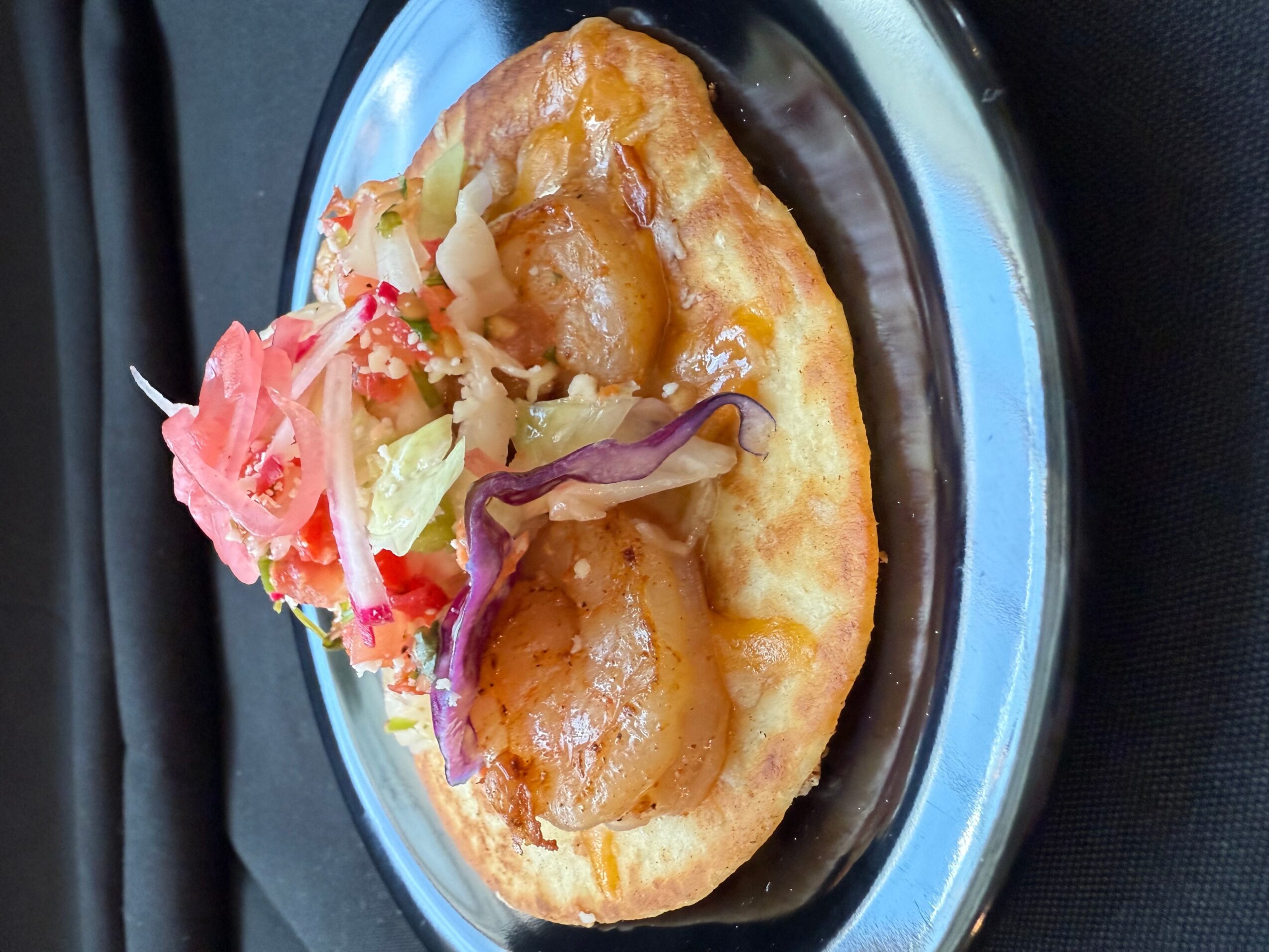 Fried bread topped with shrimp, cabbage, and purple onion on a black and silver plate.