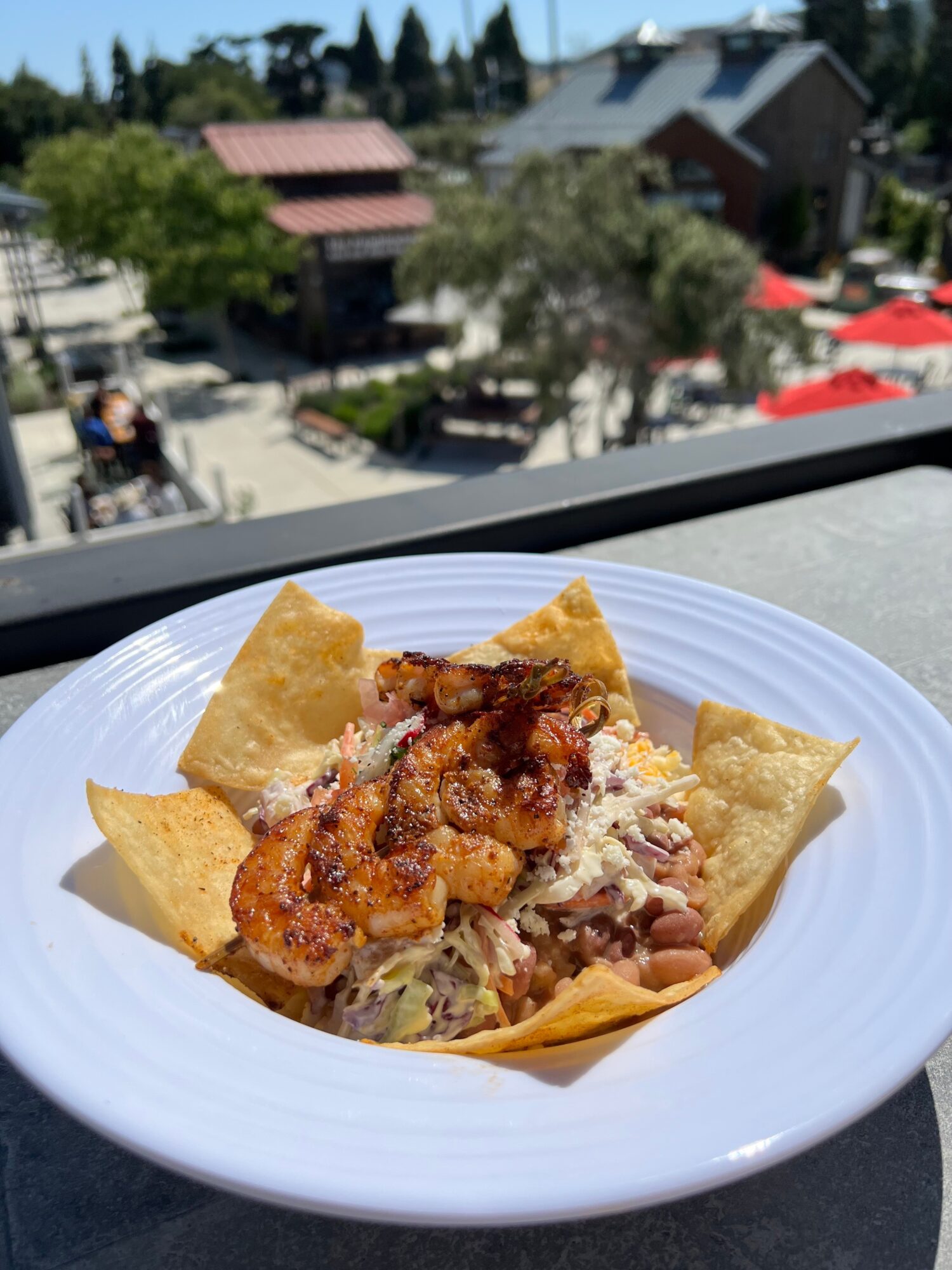 Plate with fried chicken, shredded lettuce, cheese, and tortilla chips on a table outdoors, with buildings and trees in background.