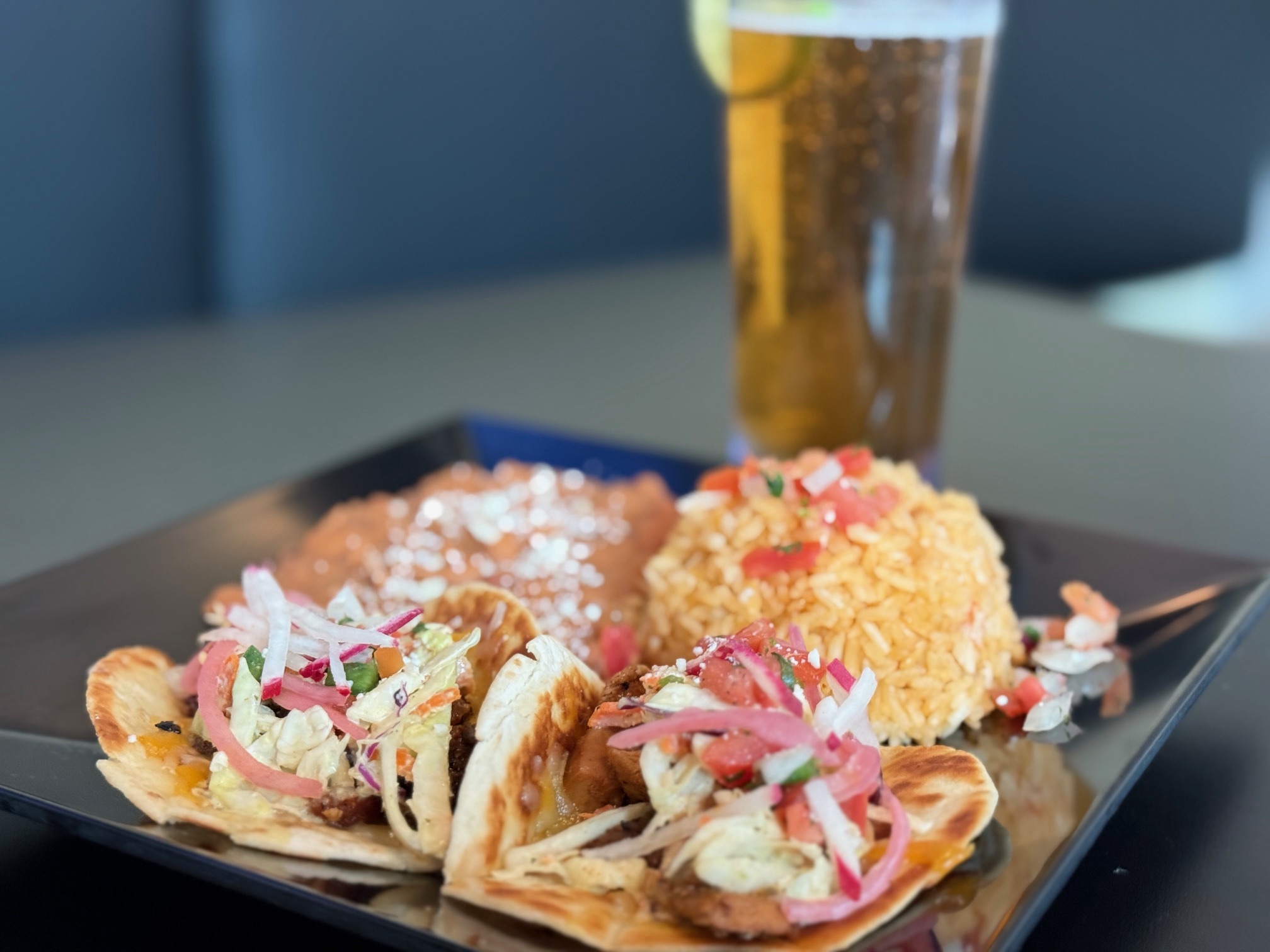 Three tacos with shredded meat, onions, and herbs on a black plate, with a glass of beer in the background.