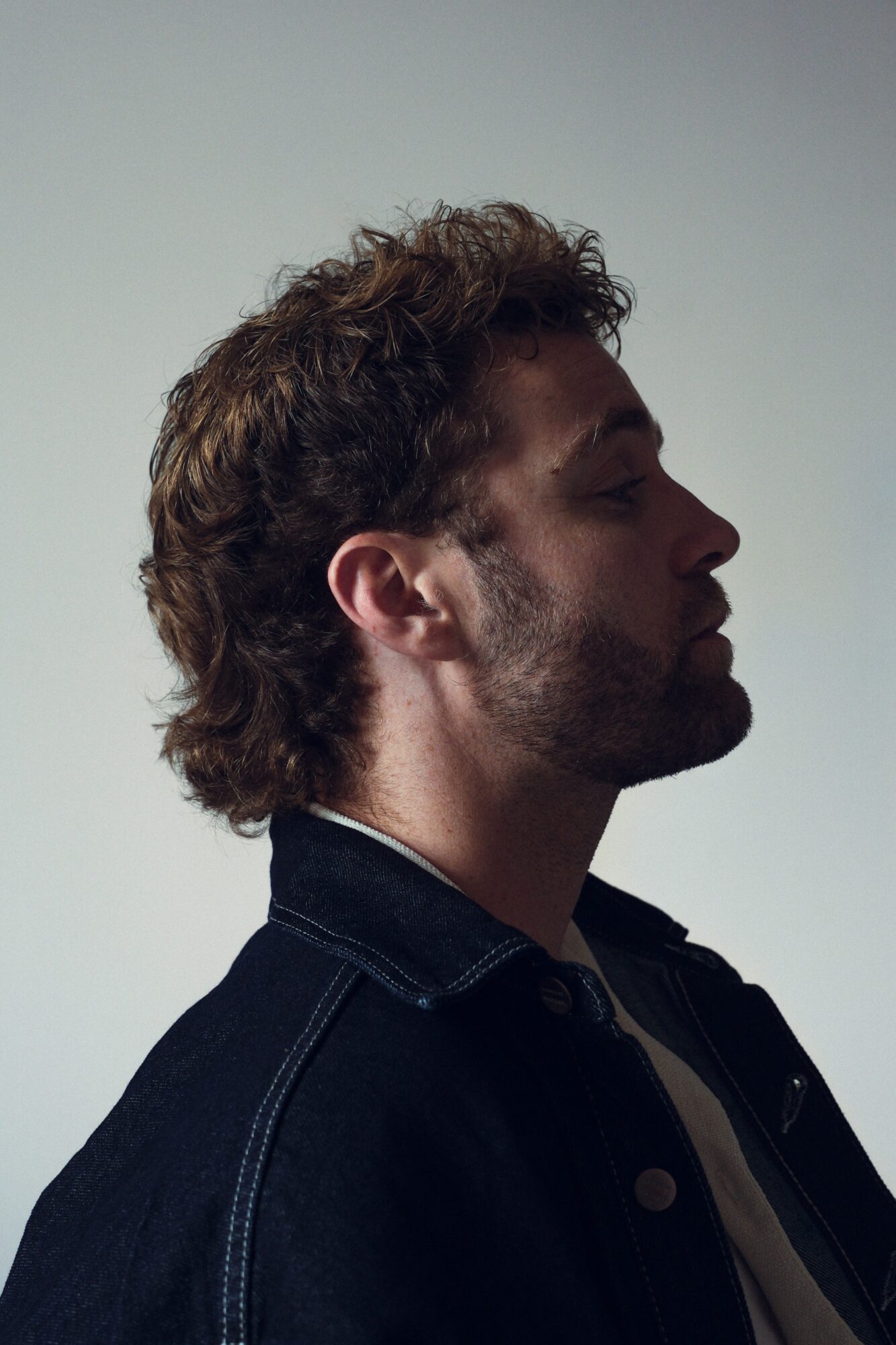 Side profile of a man with curly hair and beard, wearing a dark jacket, against a plain background.