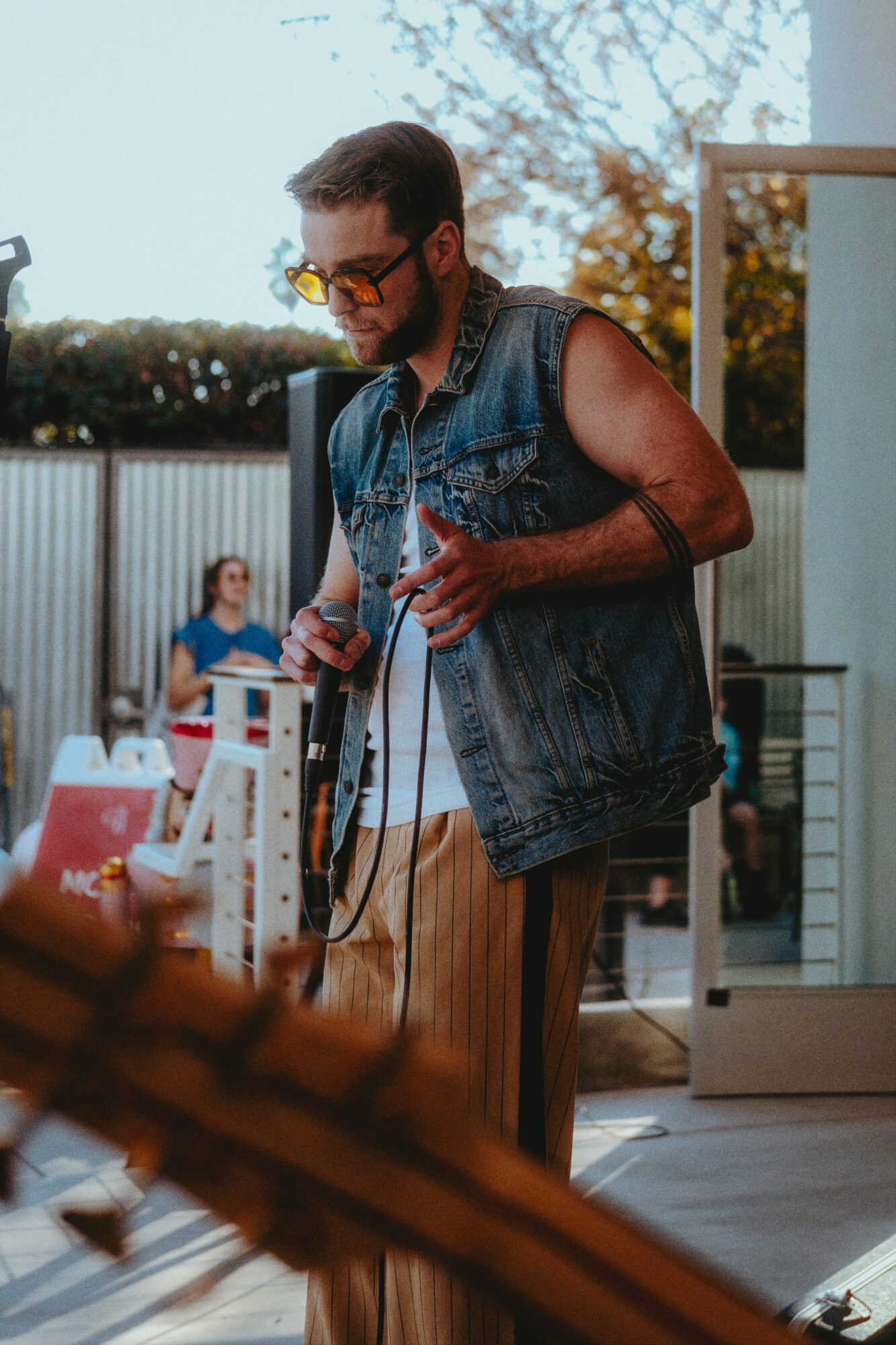 Man with sunglasses and a denim vest holding a microphone outdoors, with people and trees in background.