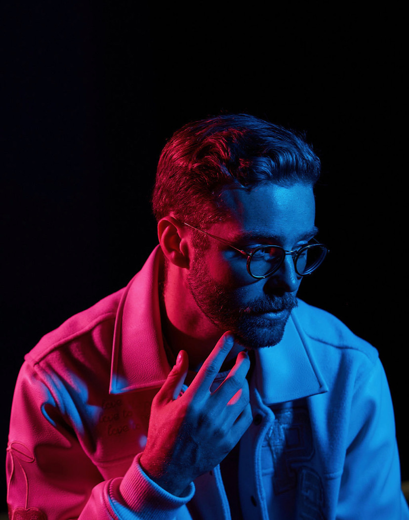 Man with glasses and beard, touching chin, illuminated by pink and blue lighting, dark background.