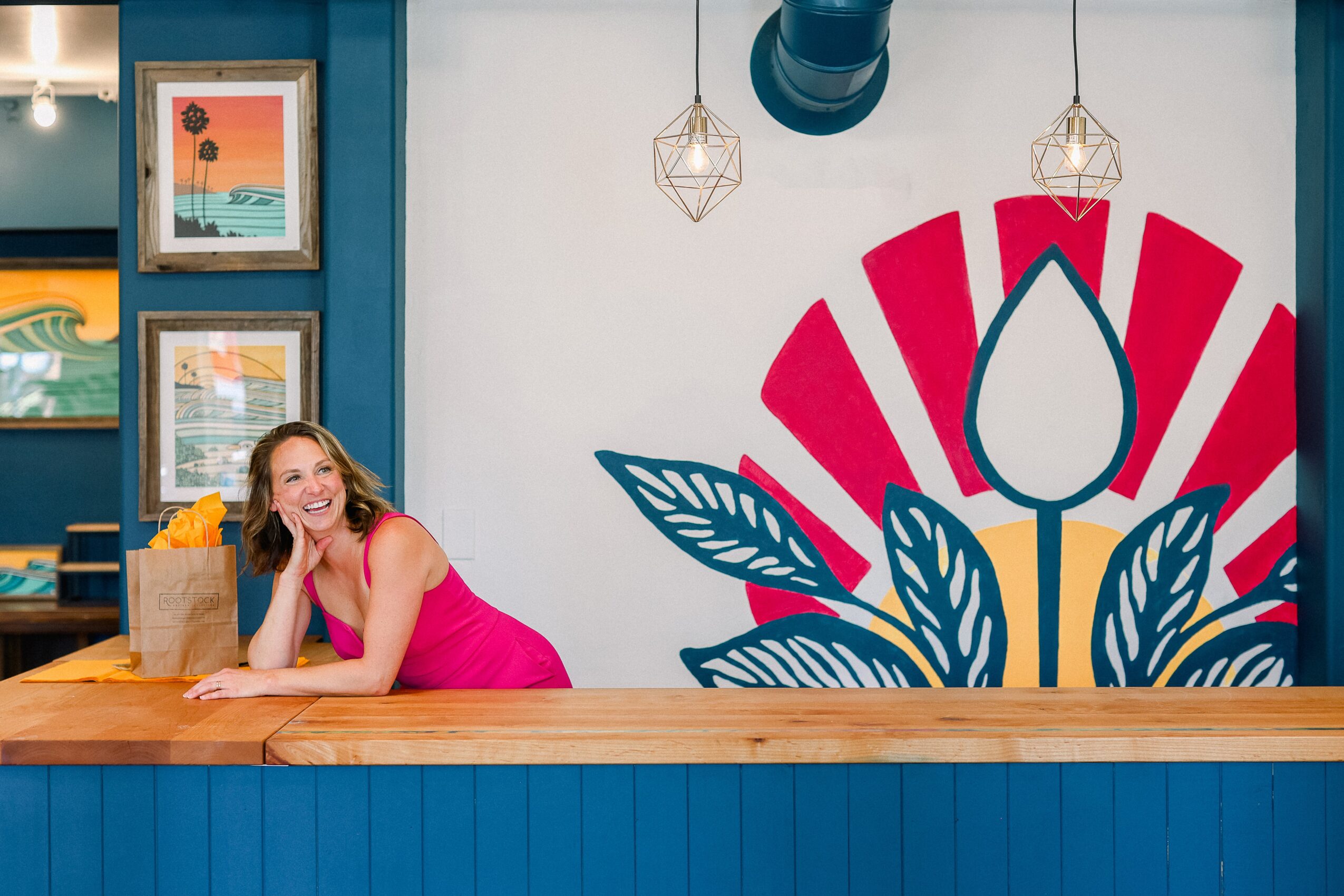 Woman in pink dress leaning on a wooden counter, smiling, with colorful wall art and a large floral mural behind her.