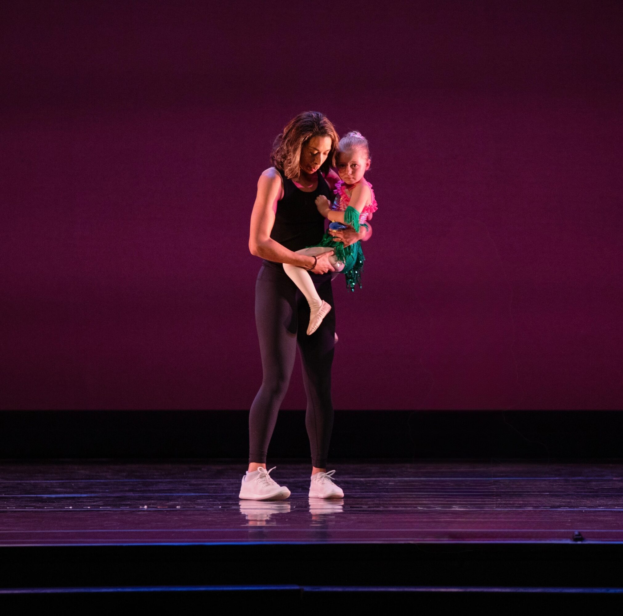 A woman holding a young girl on stage with a dark pink background, both facing forward, the woman looking down at the girl.