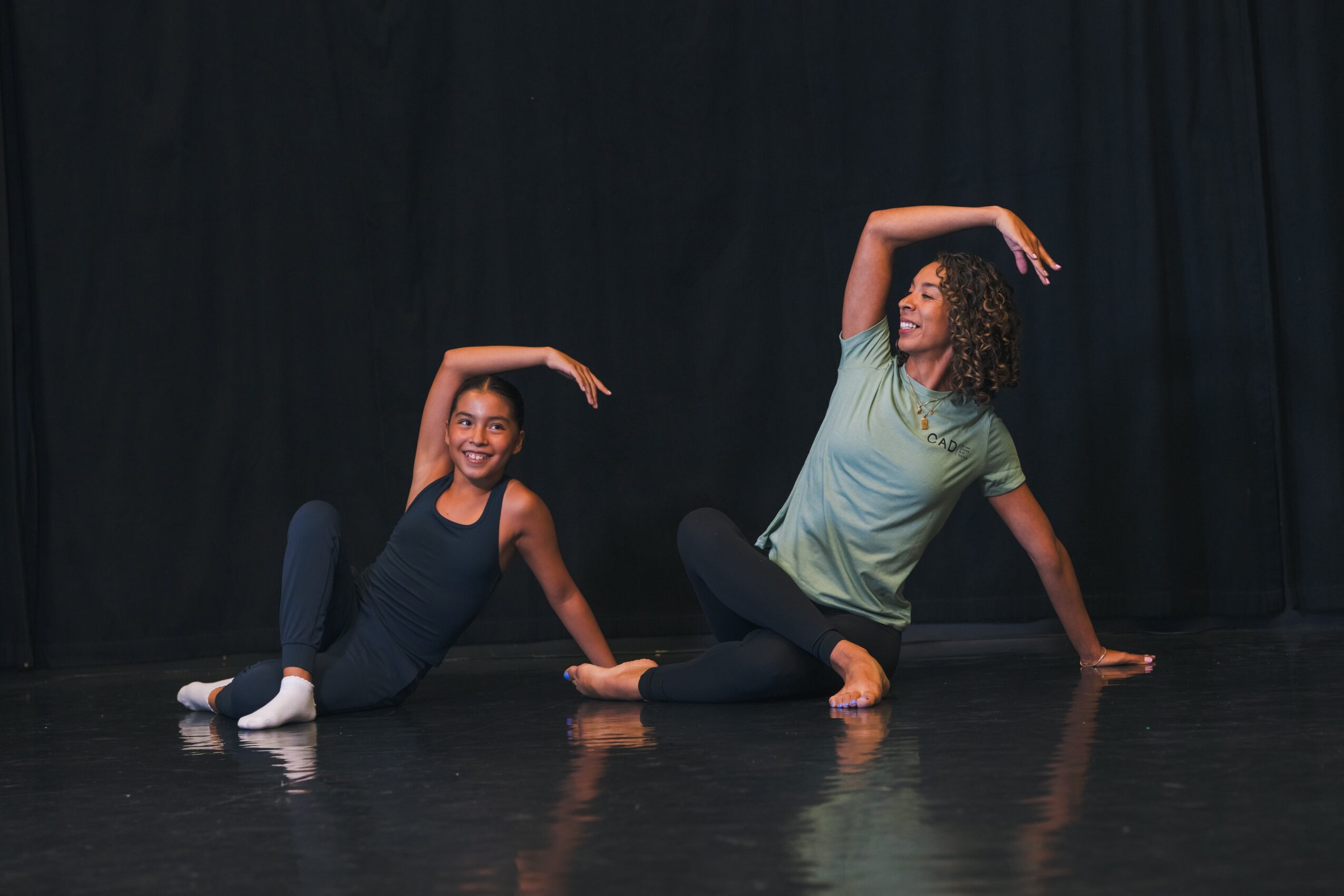 Two women sit on the floor, smiling, with one arm raised above their heads, against a black curtain background.