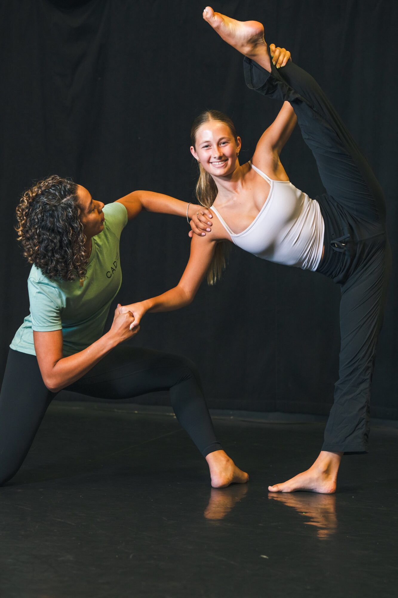 Two women practicing dance or yoga, one balancing on one leg with an extended arm, the other supporting and smiling.