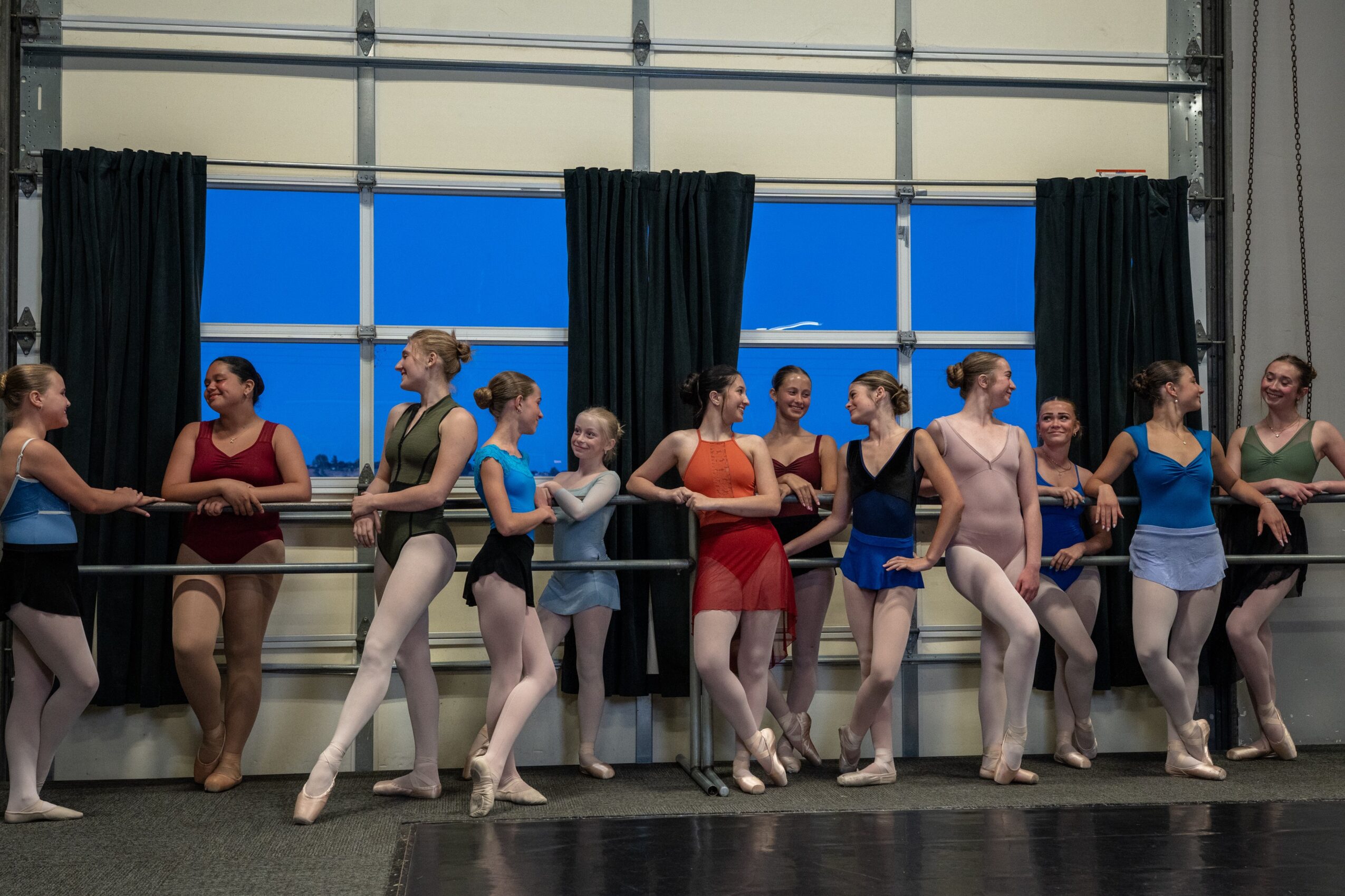 Group of ballet dancers in practice, holding a barre, wearing leotards and tights, in a dance studio with large windows and curtains.