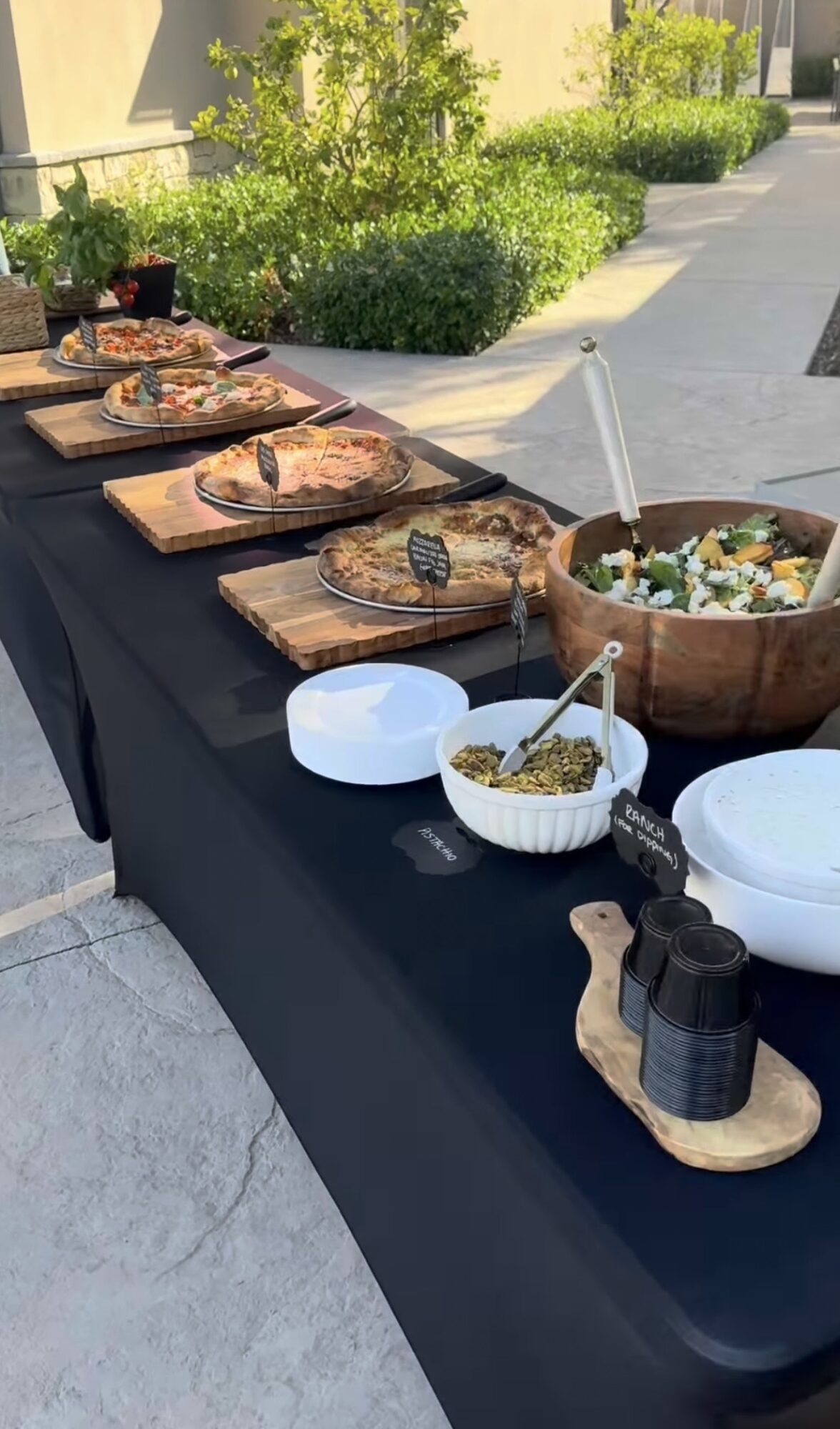 Outdoor table with various dishes, bowls, and utensils on a black tablecloth, with greenery and sidewalk in background.