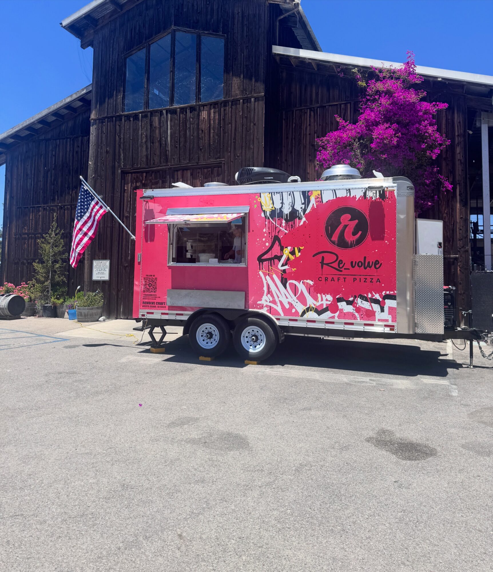 Pink food truck with graffiti-style design parked in front of a wooden building with a purple flowering tree, American flag nearby.
