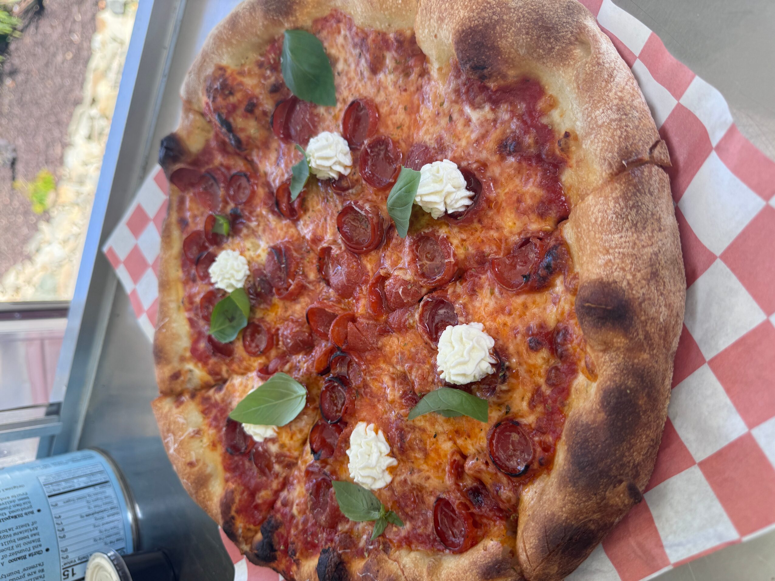 Pizza with pepperoni, cheese, and basil leaves on checkered paper, viewed from above.