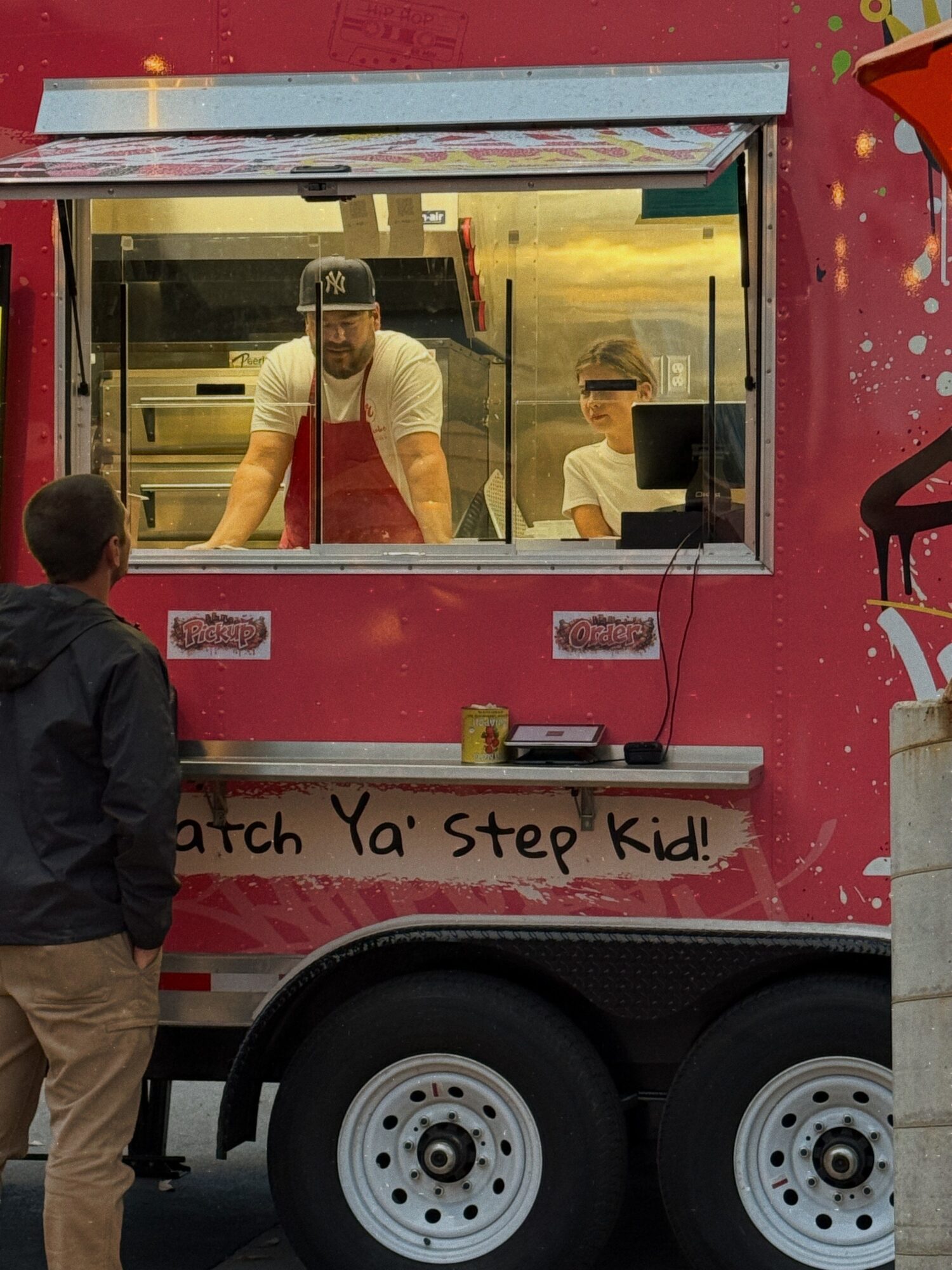Person inside a food truck serving a child through a window, with a man outside watching, pink truck with writing, and a wheel visible.