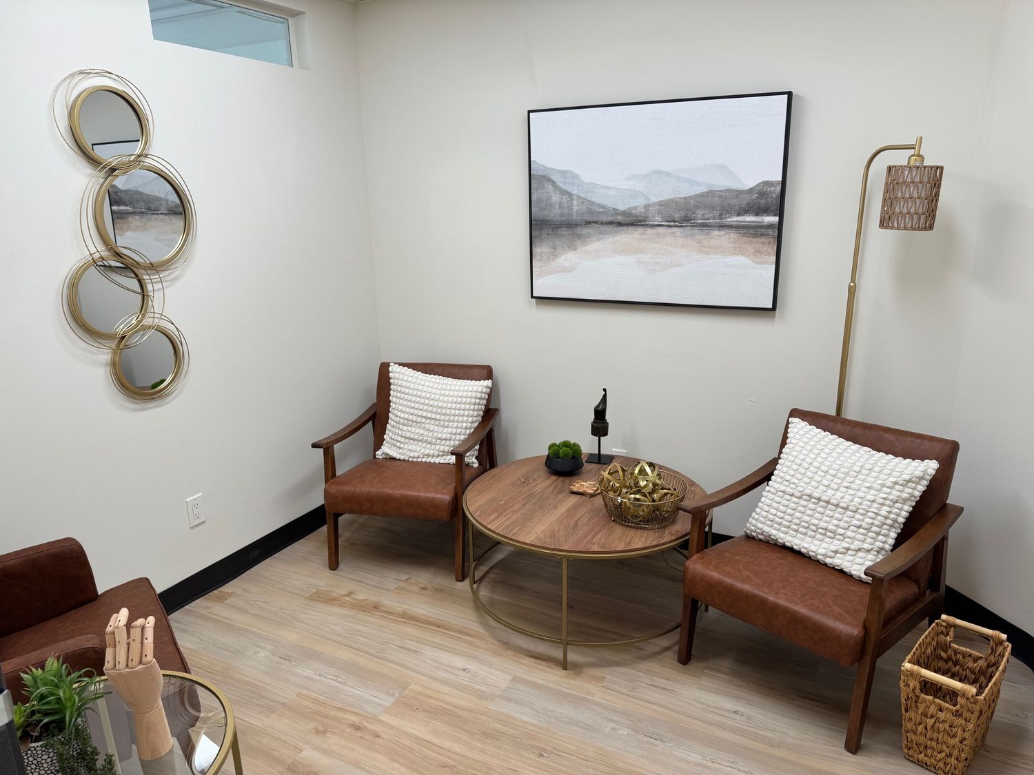 Living room with two wooden chairs, a round table, wall art, and decorative mirrors, with light-colored walls and wooden flooring.