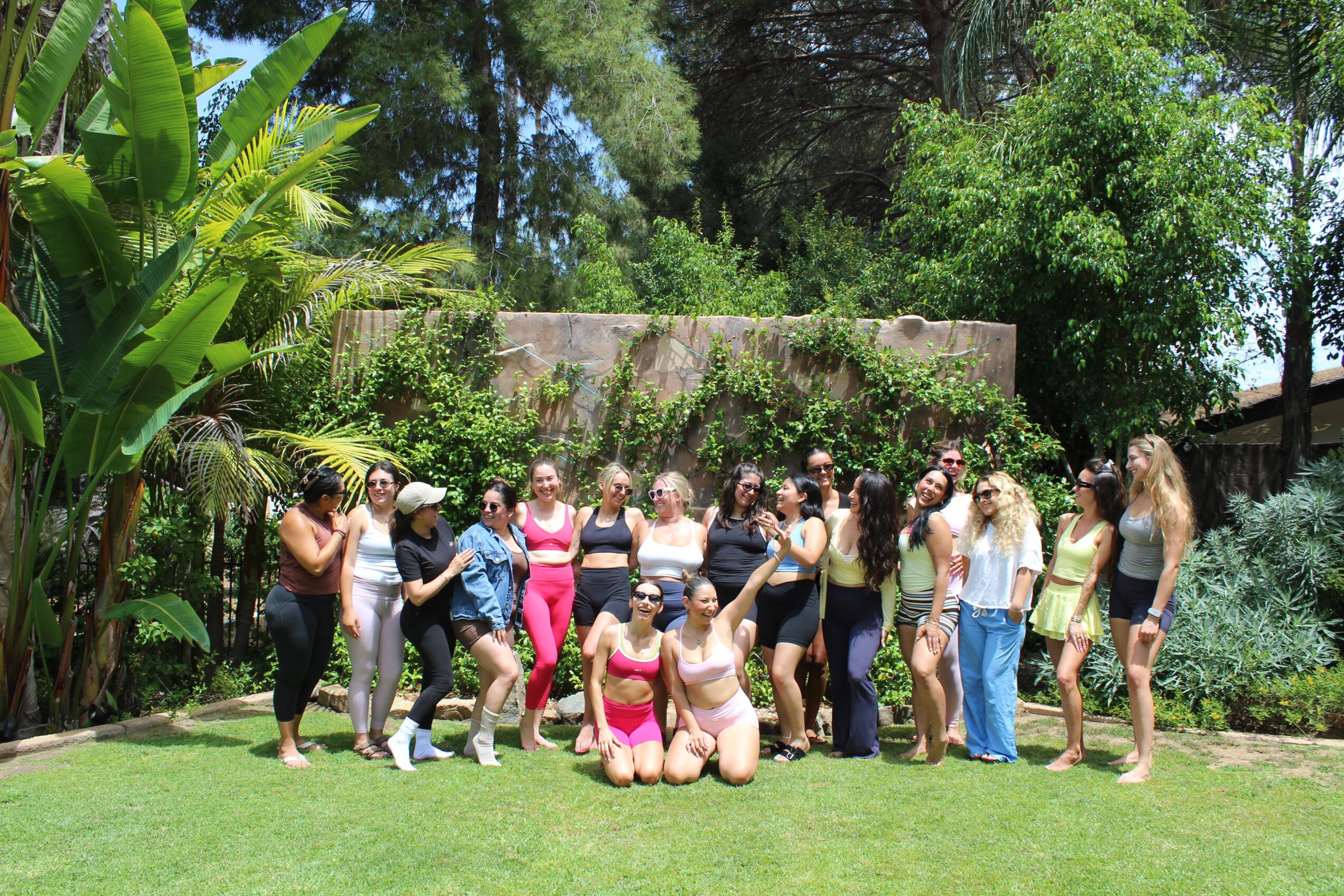 Group of people outdoors on grass with trees and bushes in background, some standing and some kneeling, smiling and posing.