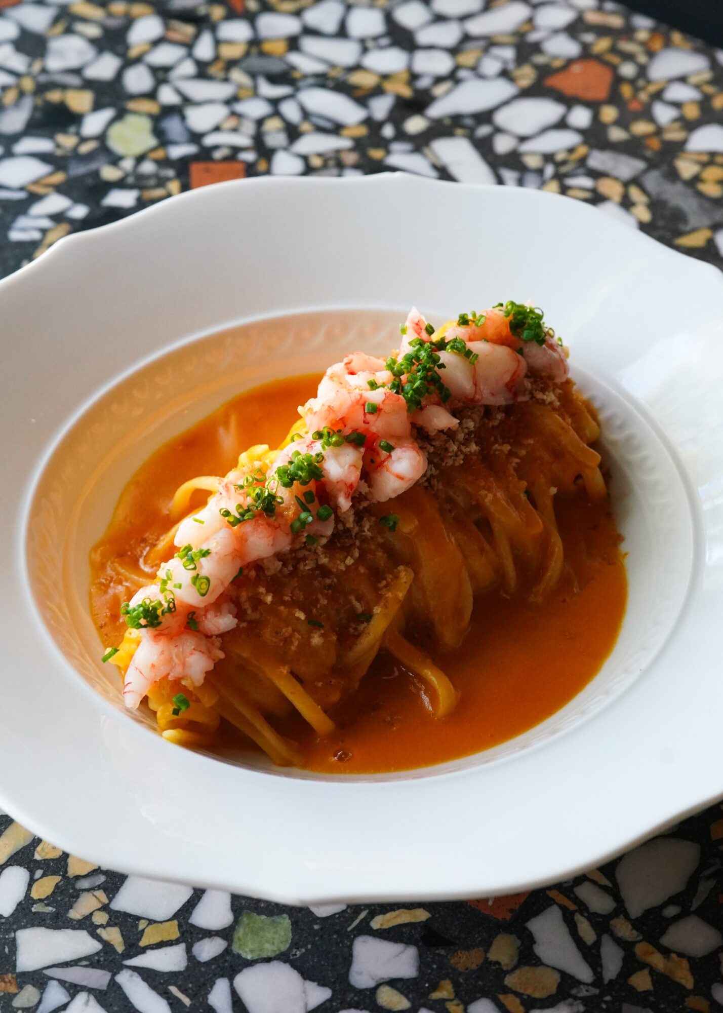 Plate of pasta with sauce, topped with pinkish seafood and green herbs, on a patterned tablecloth.