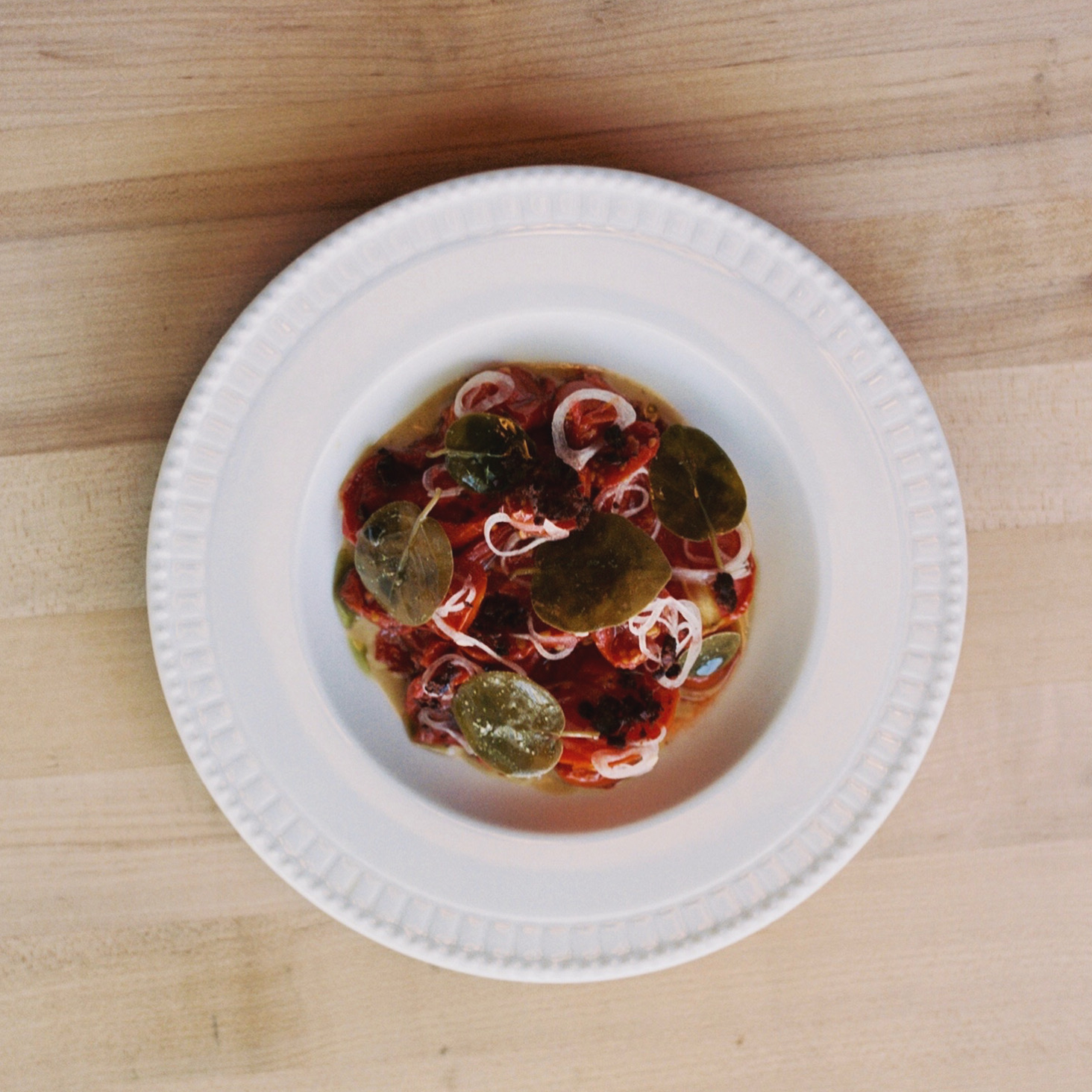 Round white plate with a dish containing green leaves, red ingredients, and white garnishes on a wooden surface.