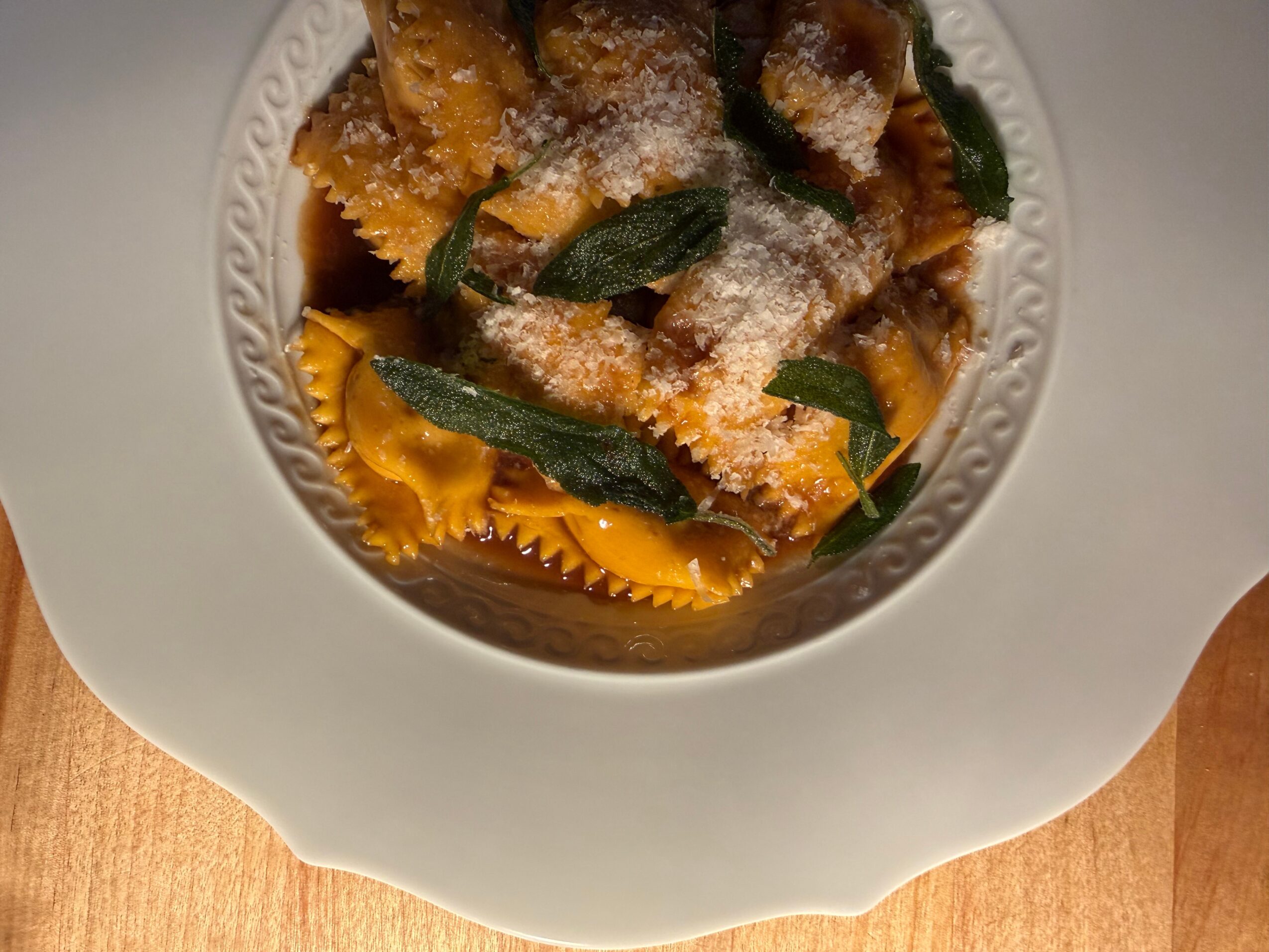 Pasta dish with fried toppings, sage leaves, and grated cheese in a white bowl on a wooden surface.