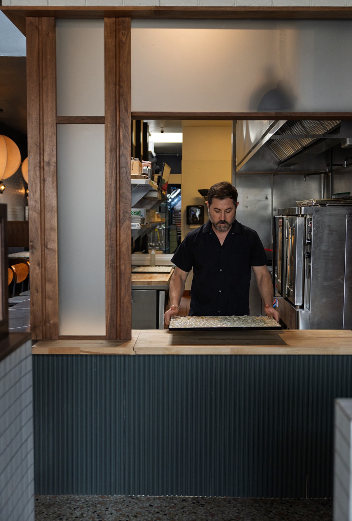 Man working behind a counter in a kitchen with stainless steel appliances, wooden framing, and a dark blue front panel.