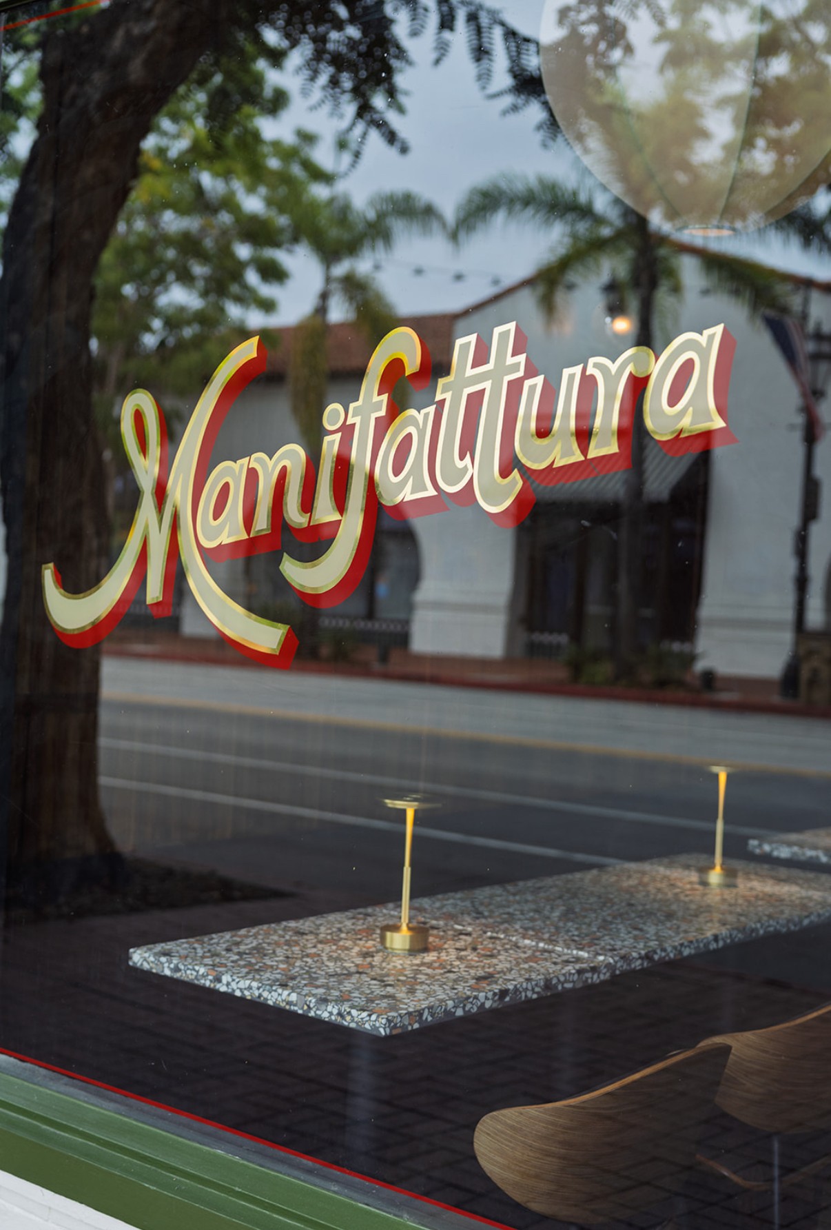 Sign with the word 'Manifattura' in red and white script, reflected in a window, with trees and buildings outside.
