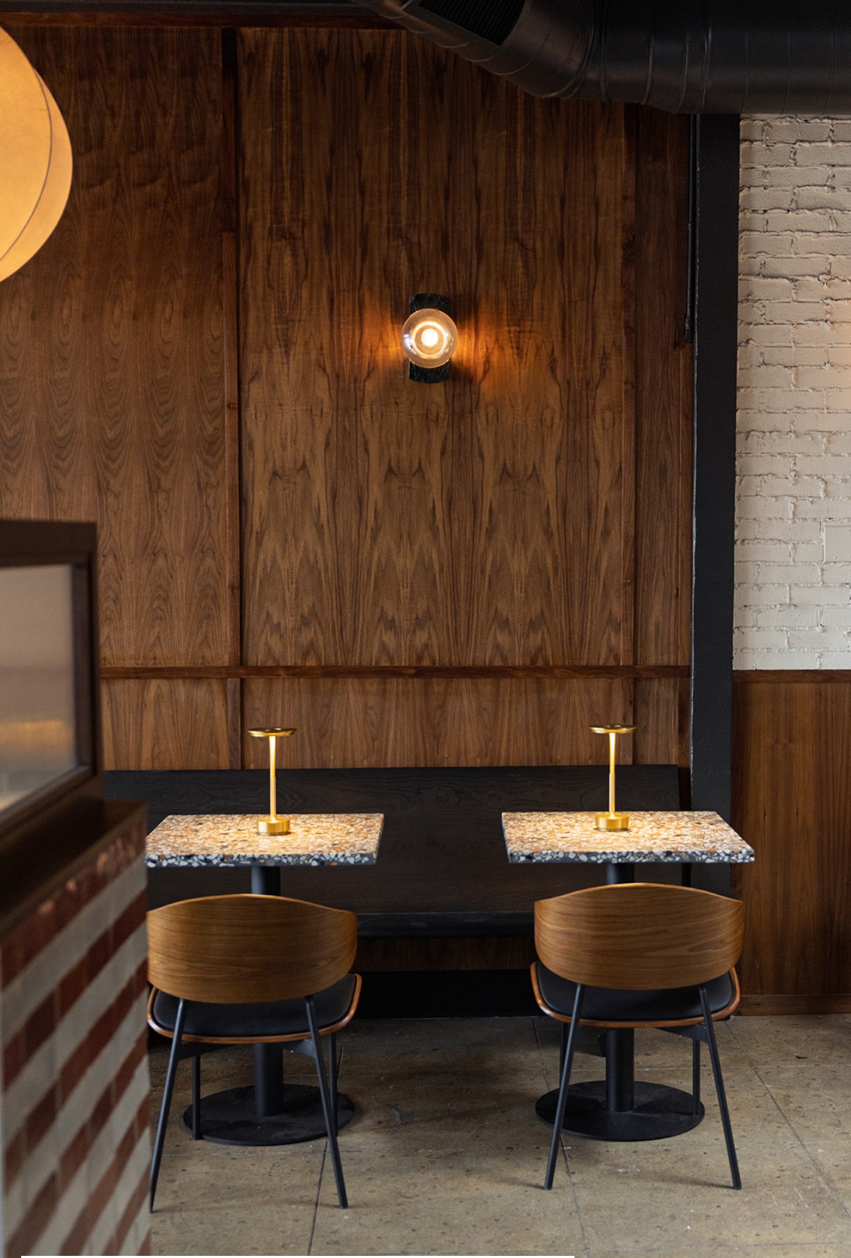 Two chairs with small tables in front, against a wooden wall with a light fixture, and a white brick wall on the right.