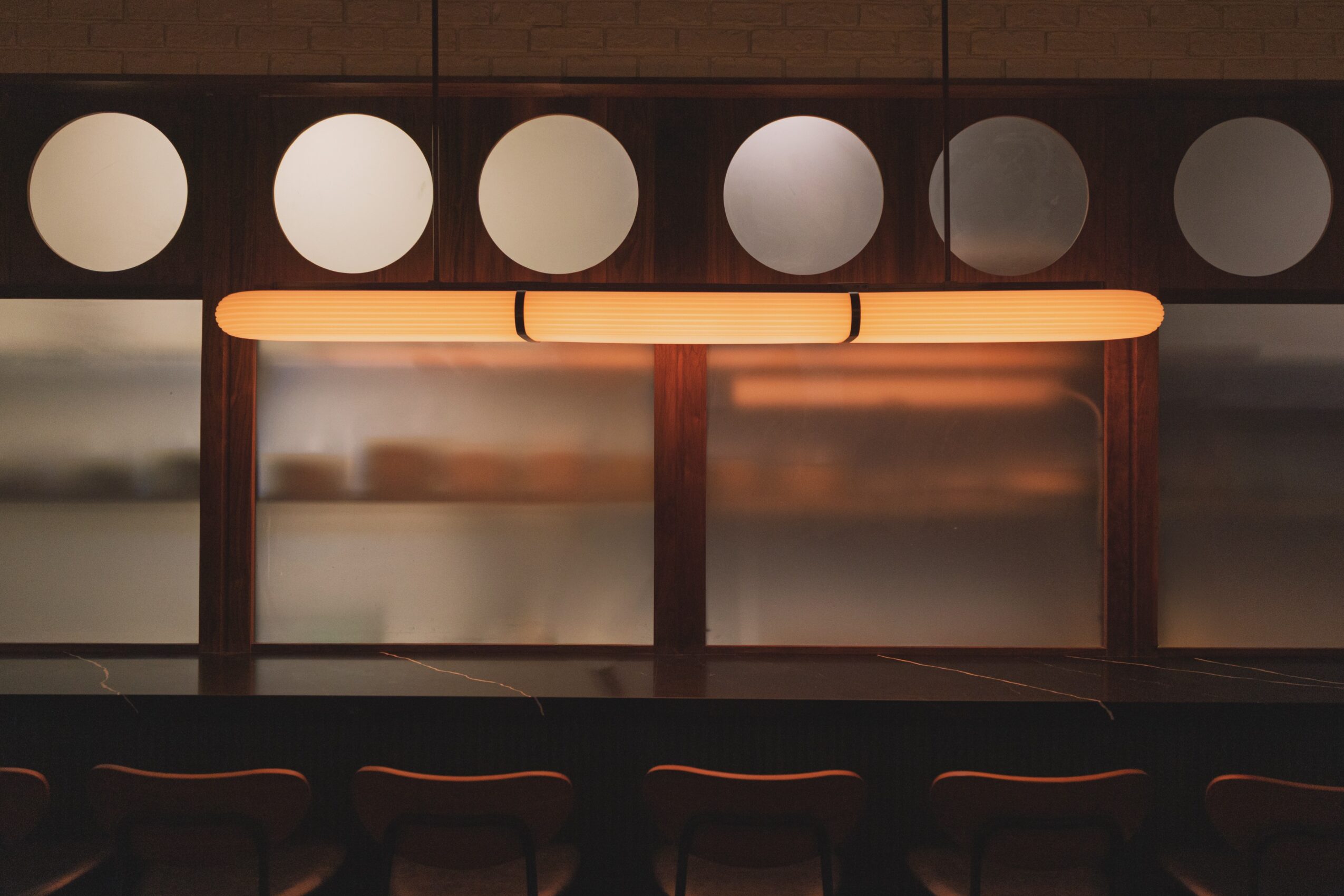 A long illuminated light fixture on a wooden table with a blurred background and circular window pattern above.