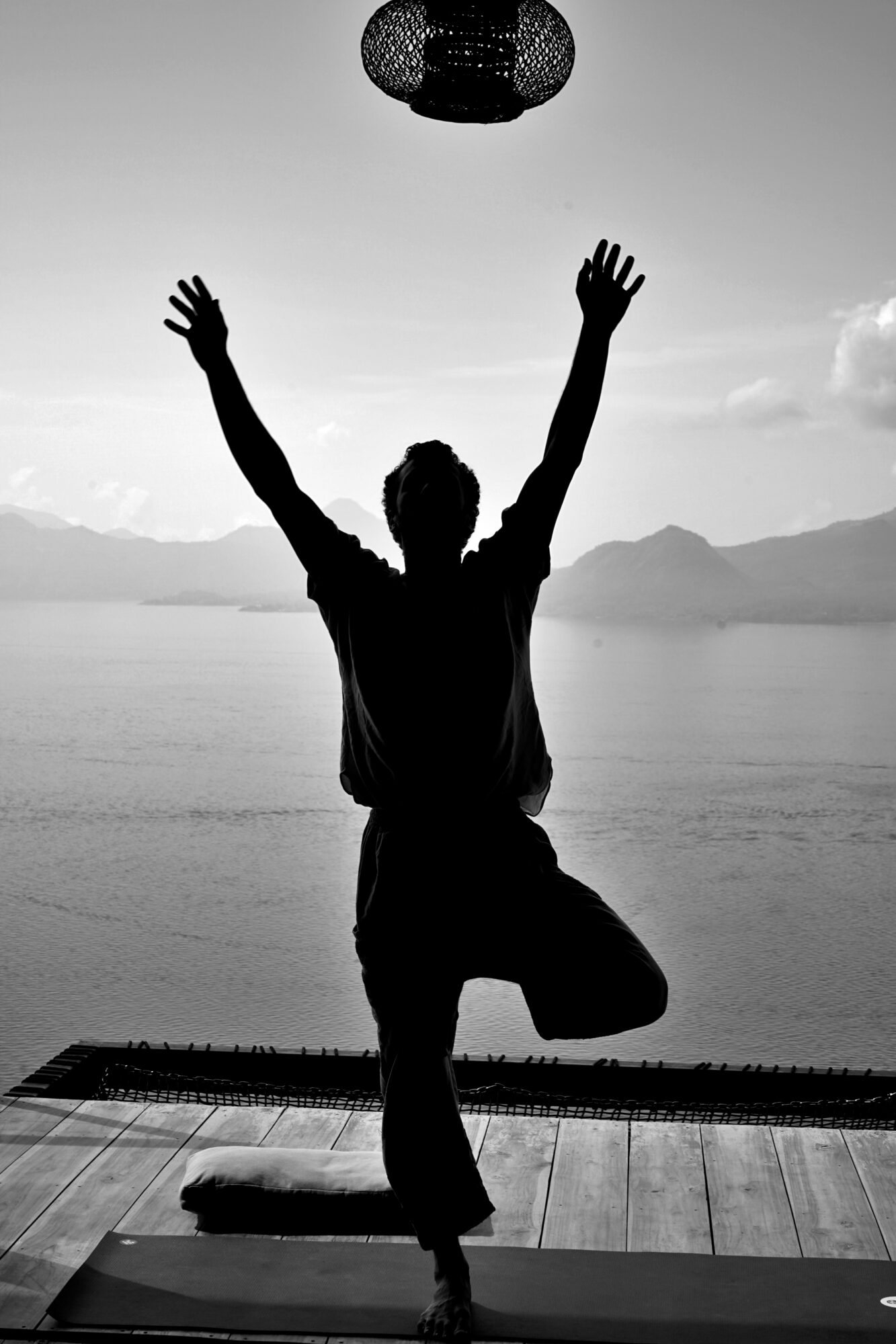 Person practicing yoga on a mat outdoors with arms raised, water and mountains in background, black and white photo.