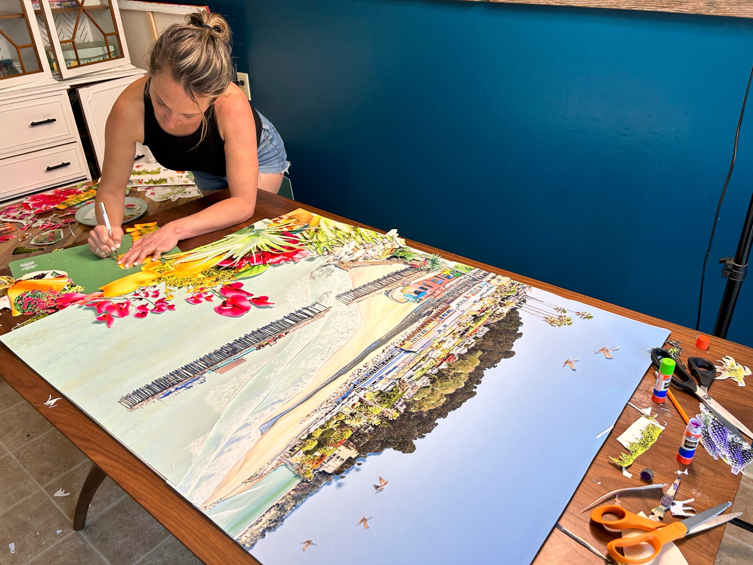 Person working on a large colorful painting on a table, with art supplies nearby, in a room with blue walls.