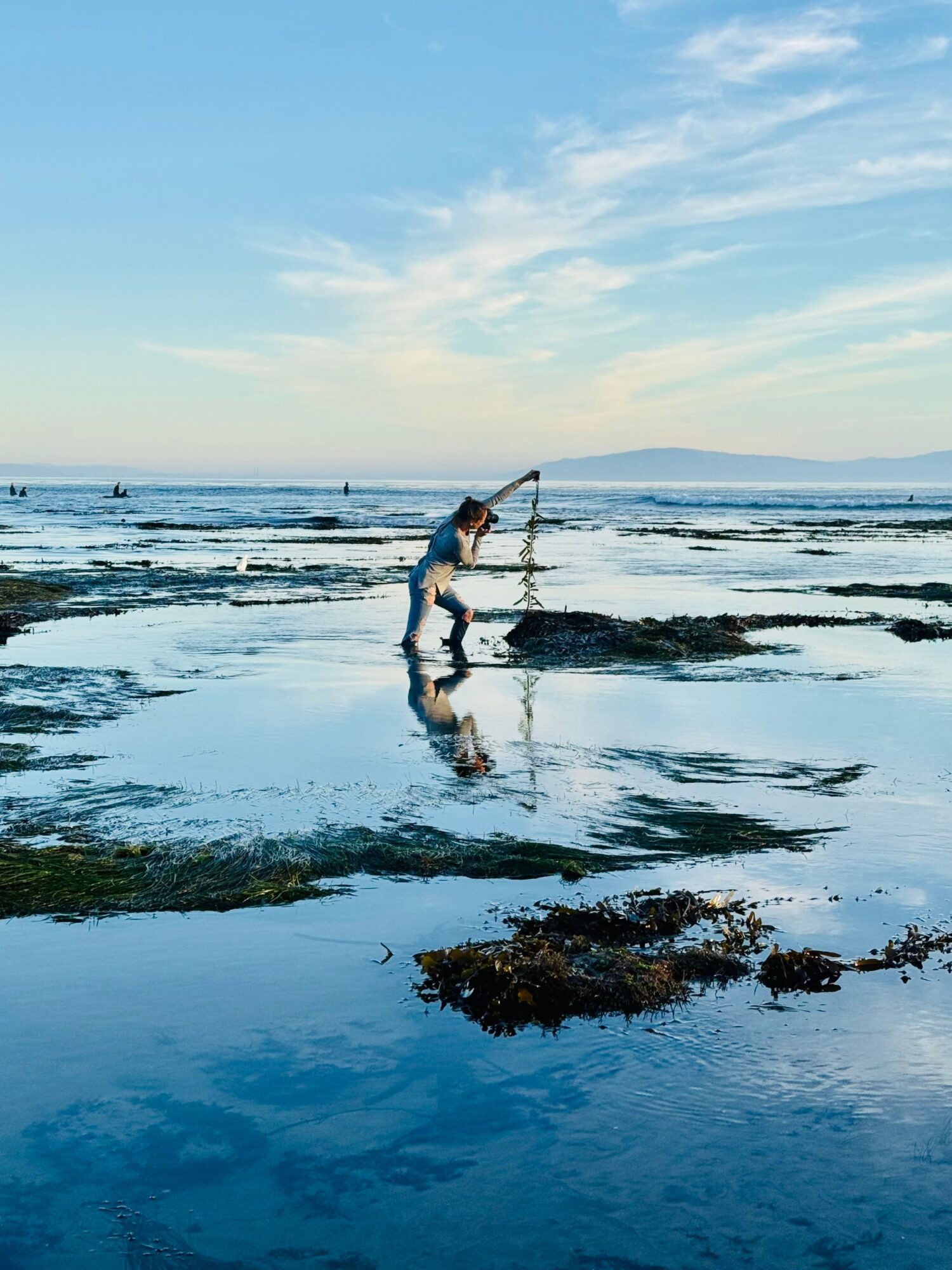 Child playing with a stick in shallow water at the beach, with rocks and a distant horizon under a blue sky.