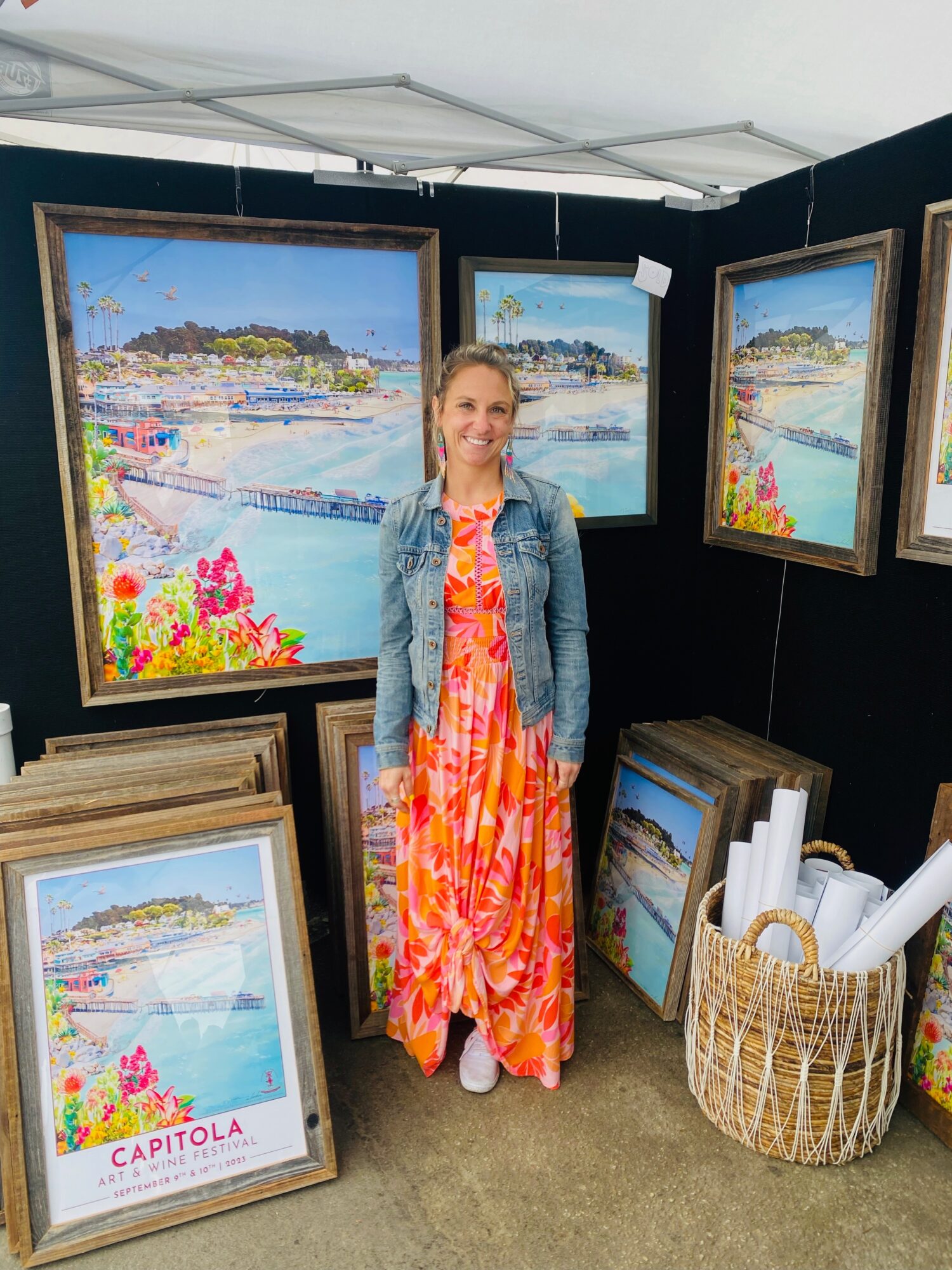 Woman in colorful dress and denim jacket standing in art display area with framed paintings and a basket of rolled papers.