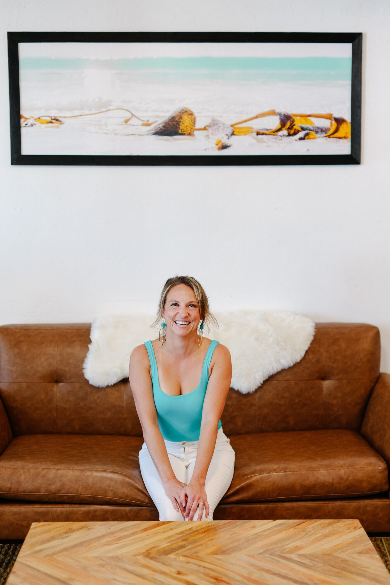 Woman sitting on a brown sofa with a white blanket, smiling, in a room with a framed landscape painting above her.
