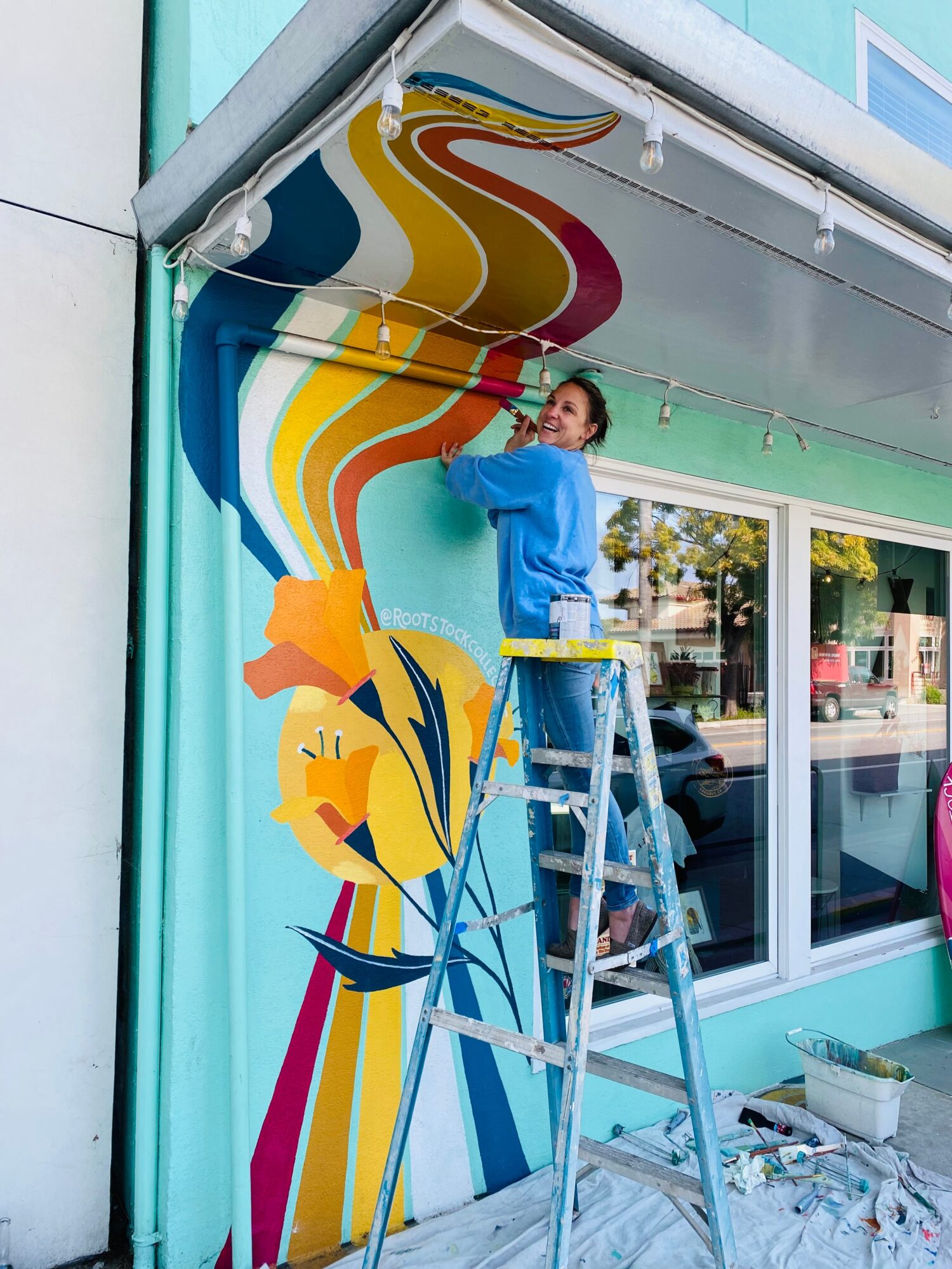 Person painting a colorful mural on a building wall, standing on a ladder, with a window and street scene in the background.