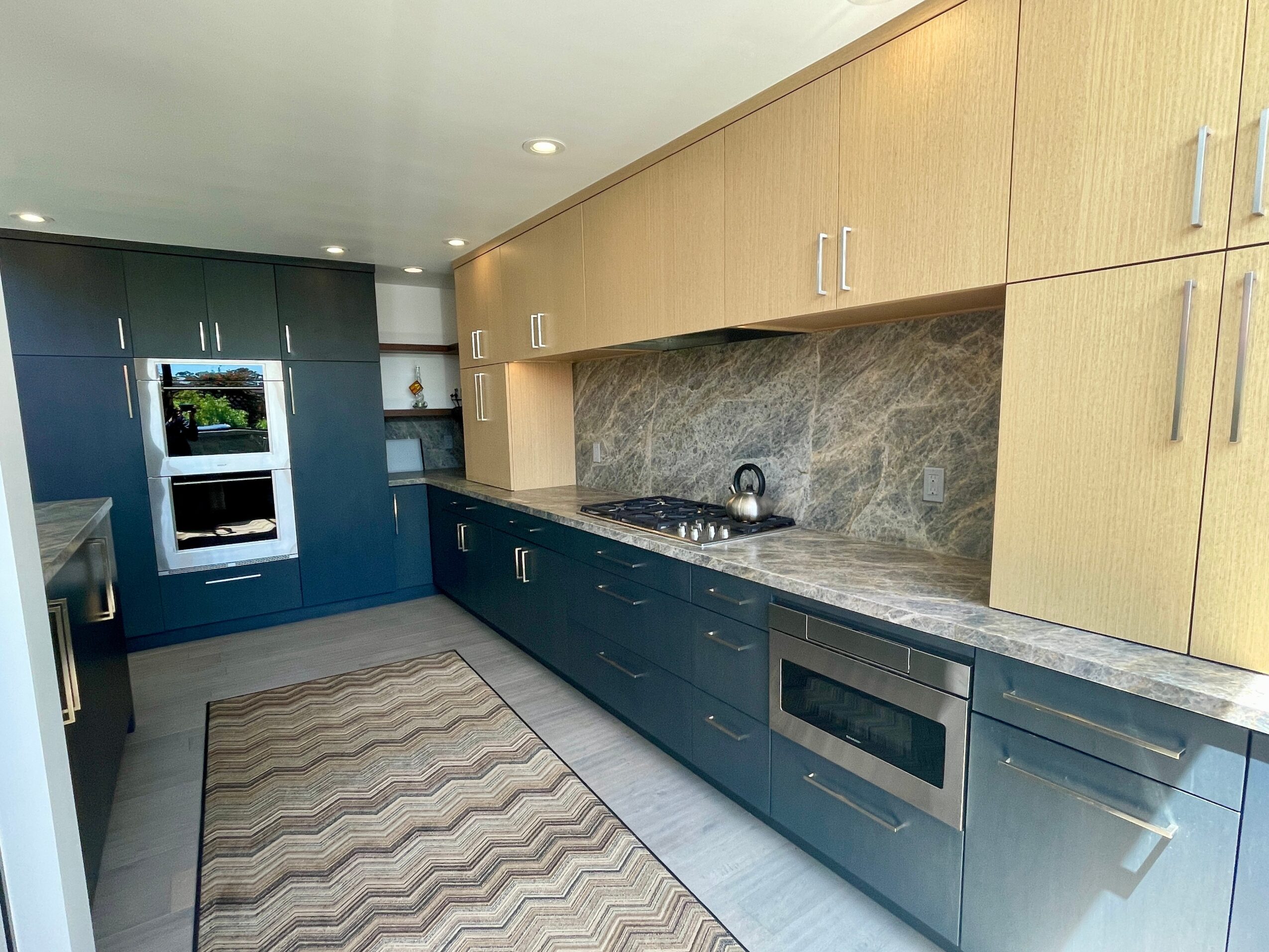 Modern kitchen with blue lower cabinets, yellow upper cabinets, marble backsplash, and built-in oven, with a patterned rug on the floor.