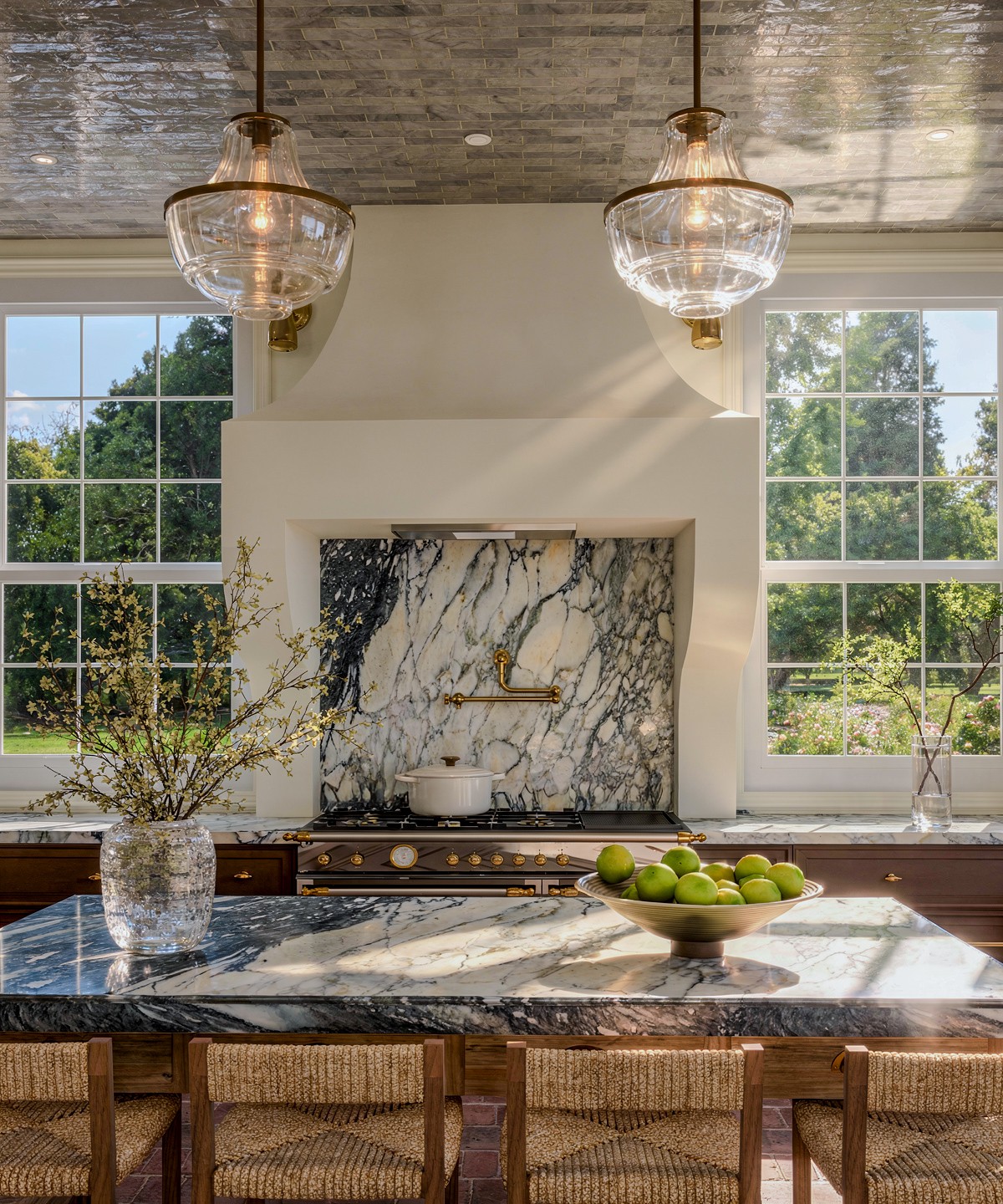 Kitchen with large windows, marble backsplash, and two hanging pendant lights above a marble countertop with a bowl of green apples.