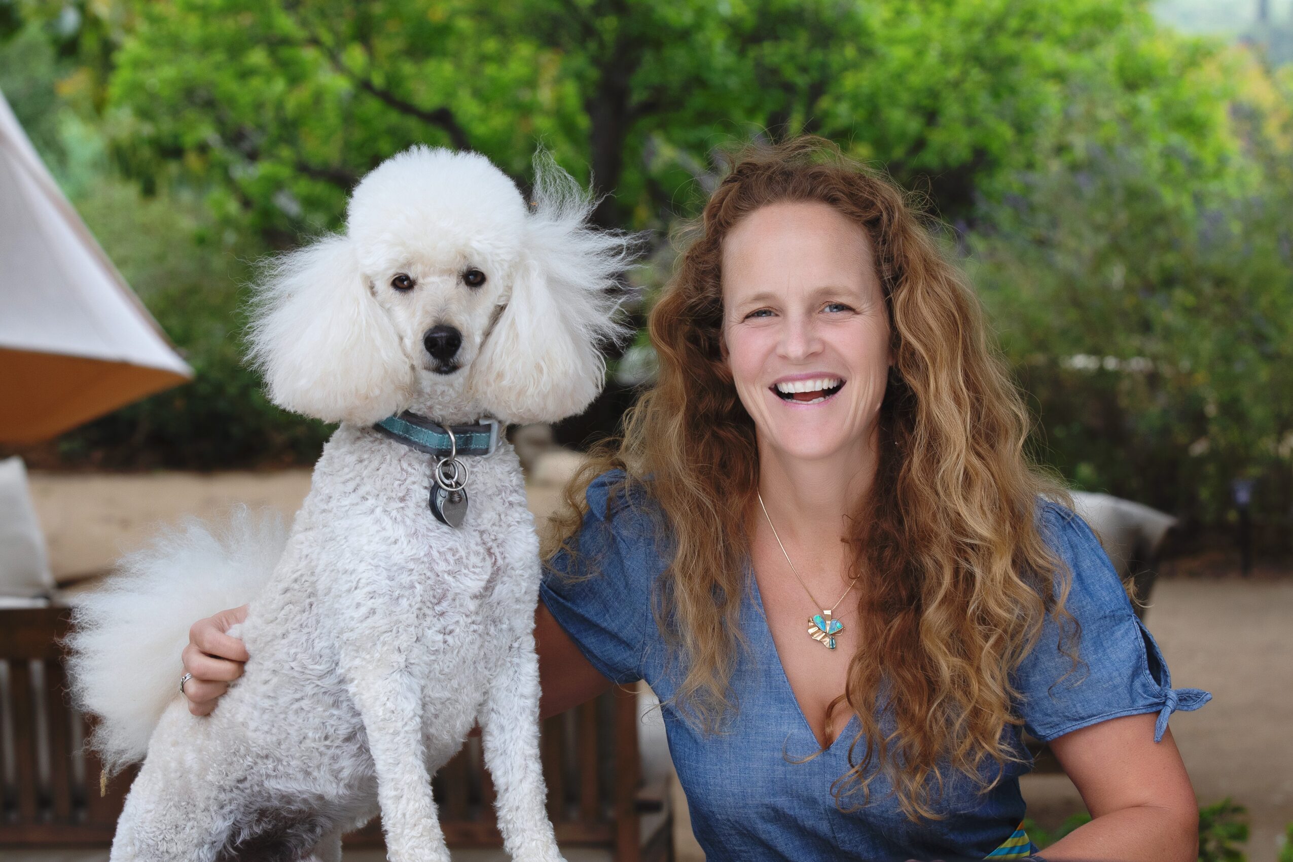 Woman with curly hair smiling outdoors next to a poodle with fluffy ears, both sitting on a bench.