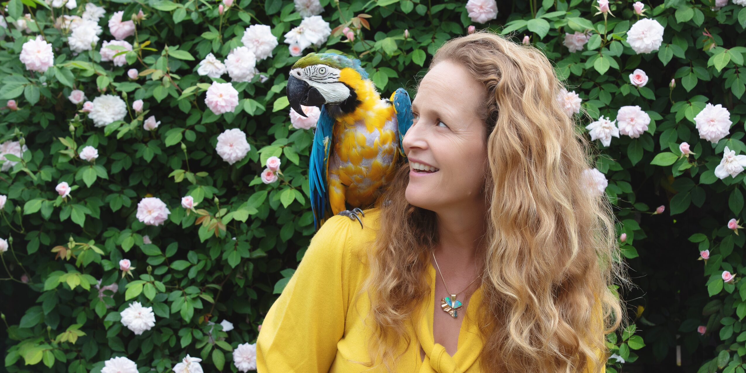 Woman with long curly hair smiling at a colorful parrot on her shoulder, surrounded by pink and white flowers.