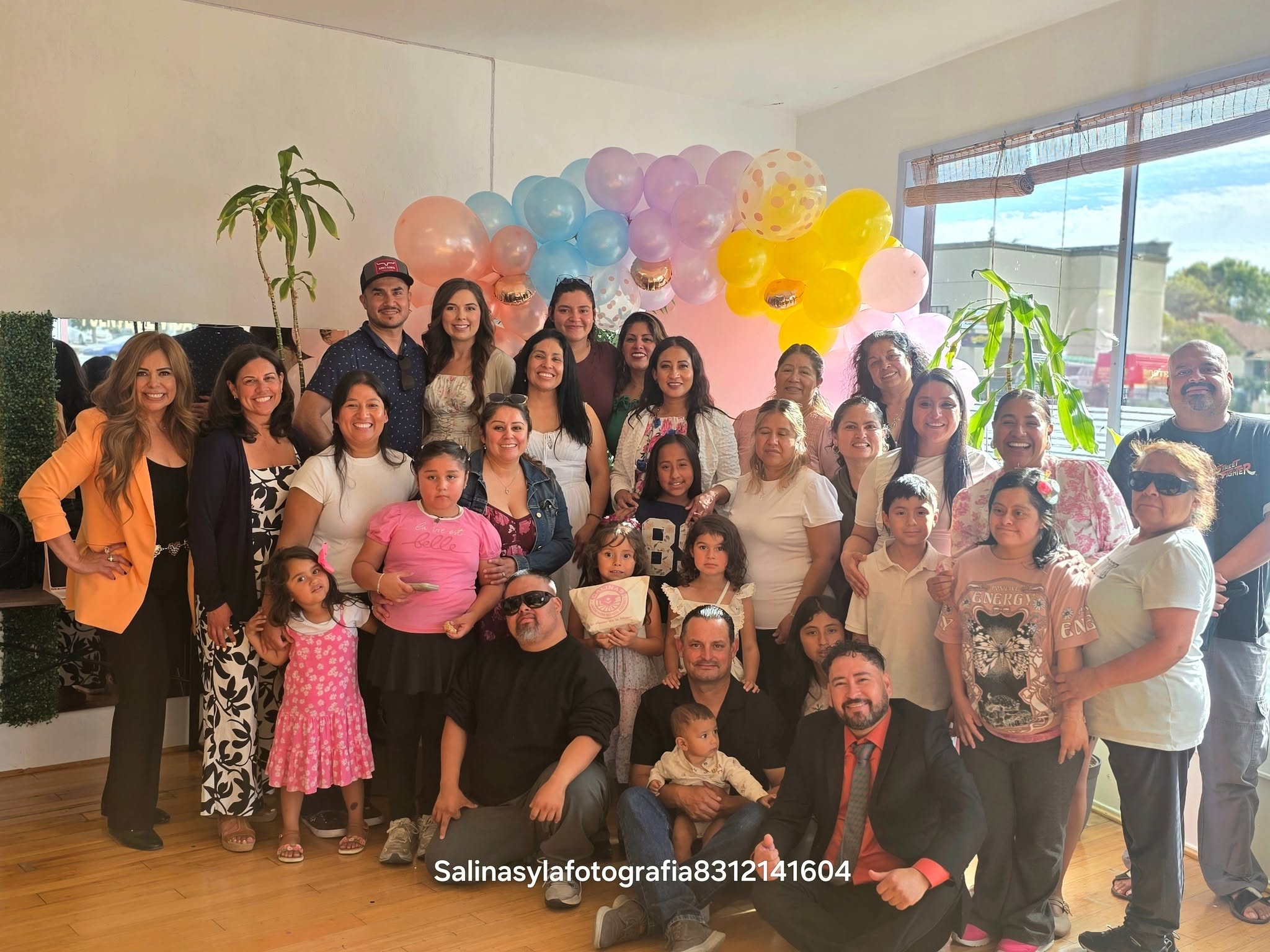 Group of people celebrating indoors with colorful balloons, some children, and a window view outside.