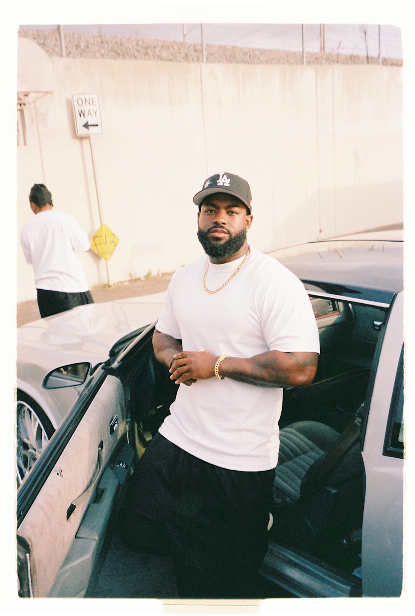 Man with beard in white t-shirt and cap leaning against a car door, with a woman in white in the background.