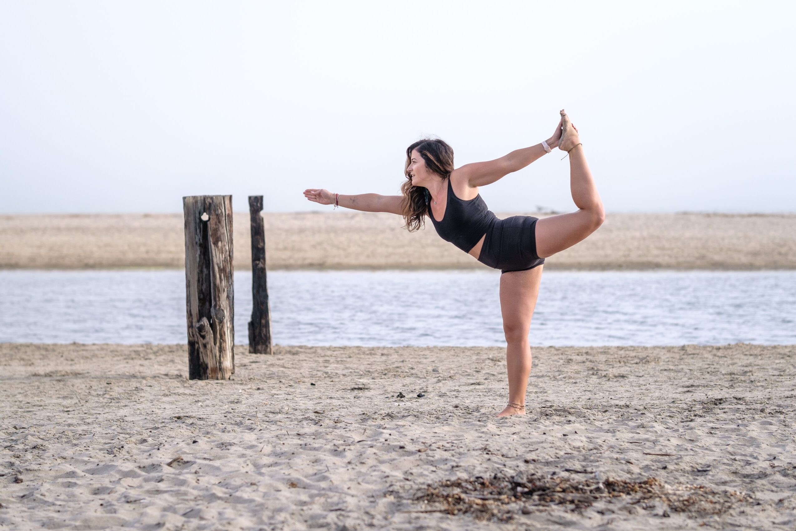 Woman practicing yoga on a beach, balancing on one leg with her other leg bent behind her, holding her foot with one hand.