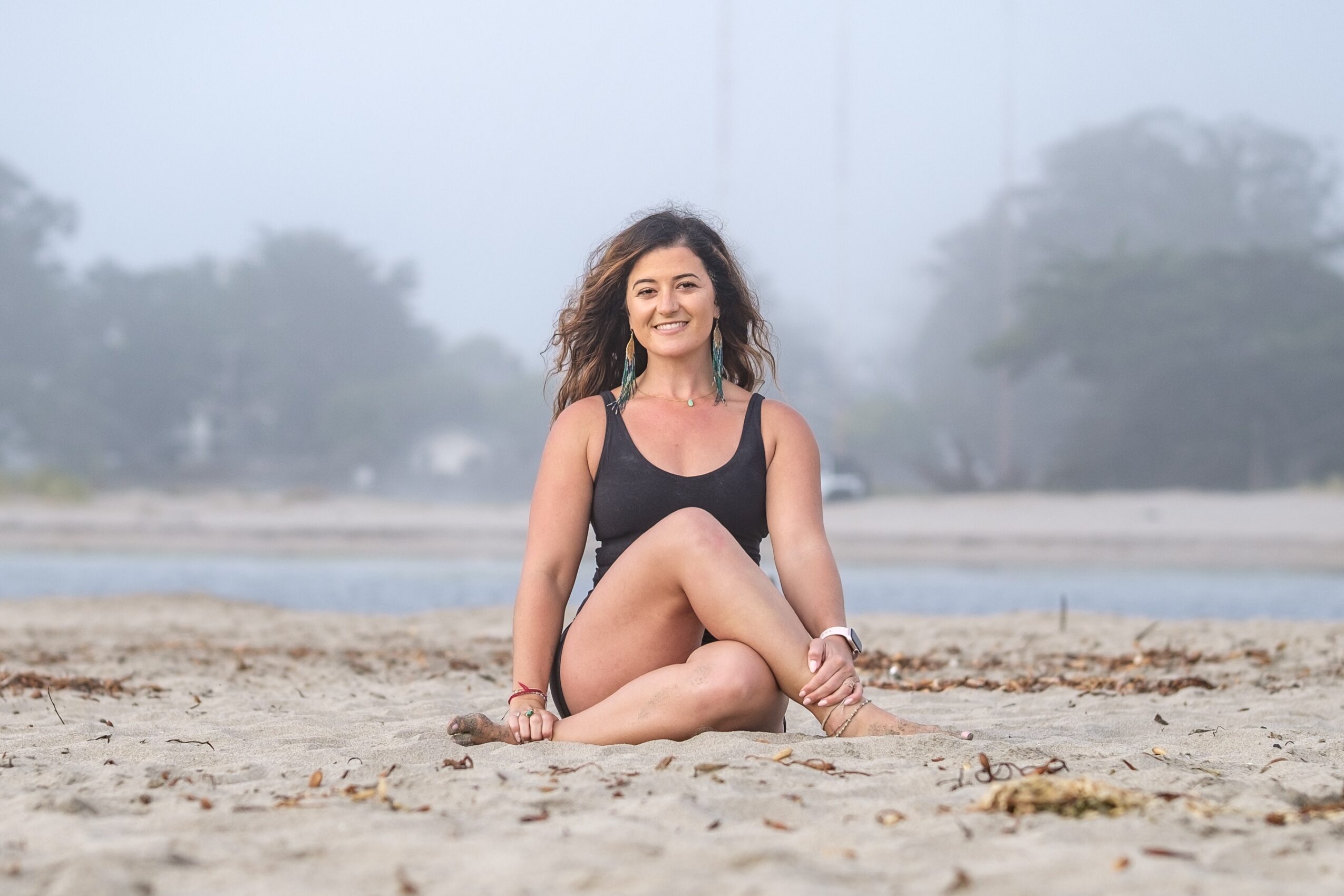 Smiling woman sitting on sandy beach with trees and fog in background, wearing a black swimsuit, legs crossed, hands on ground.