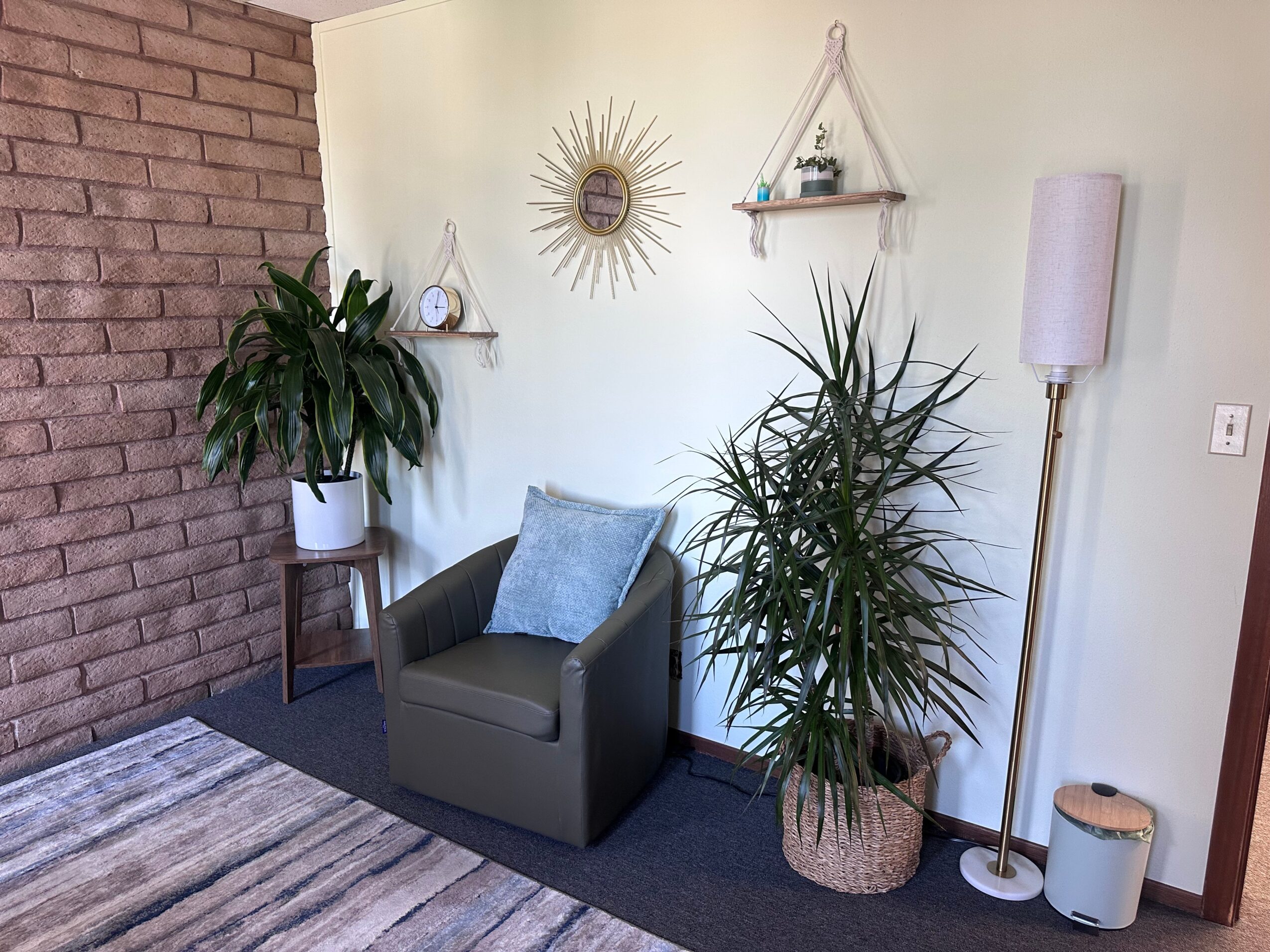Corner of a room with a gray armchair, two potted plants, a tall floor lamp, and wall decorations including a sunburst mirror.