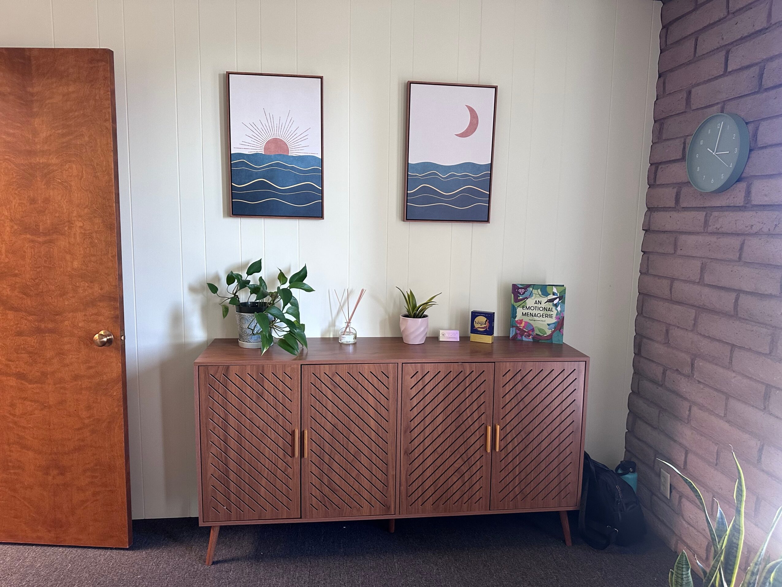Wooden sideboard with plants, candles, and decorative items, two framed moon and sun art pieces above, brick wall on right, door on left.