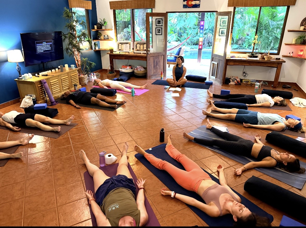 People lying on mats in a yoga or meditation class room with large windows and a person leading the session.