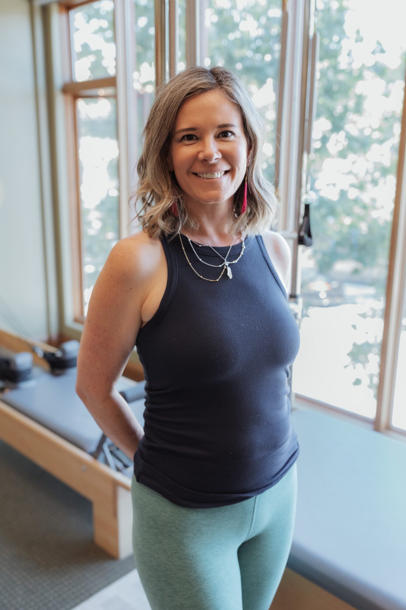 Woman with shoulder-length hair smiling, wearing a navy sleeveless top and light-colored pants, standing indoors near windows.