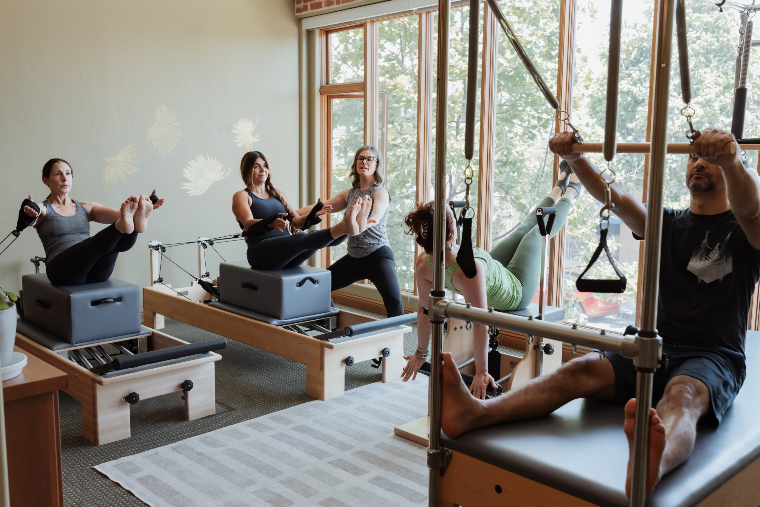 People participating in a Pilates class using reformer machines and a trapeze system in a bright studio with large windows.