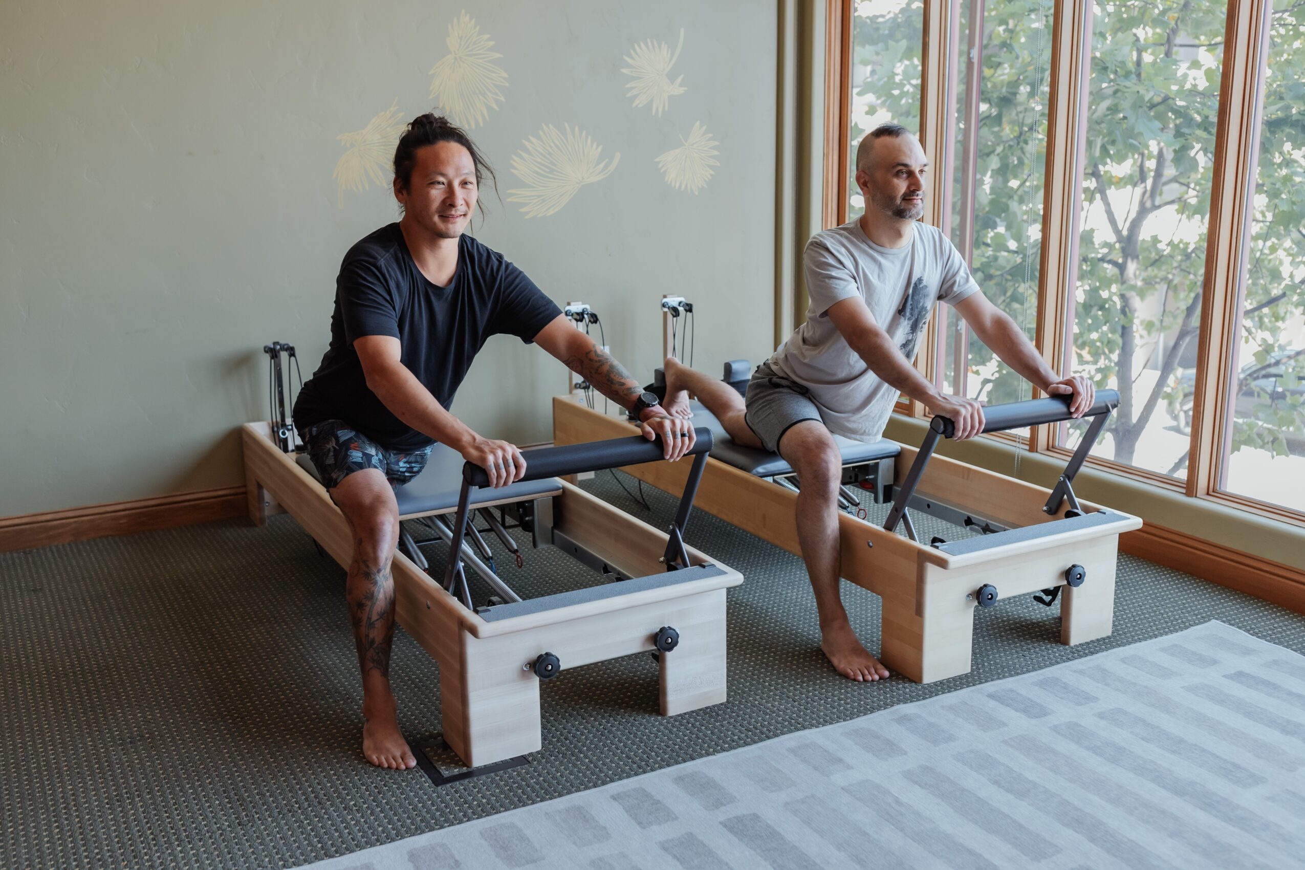 Two men using Pilates reformer machines in a room with large windows and natural light.