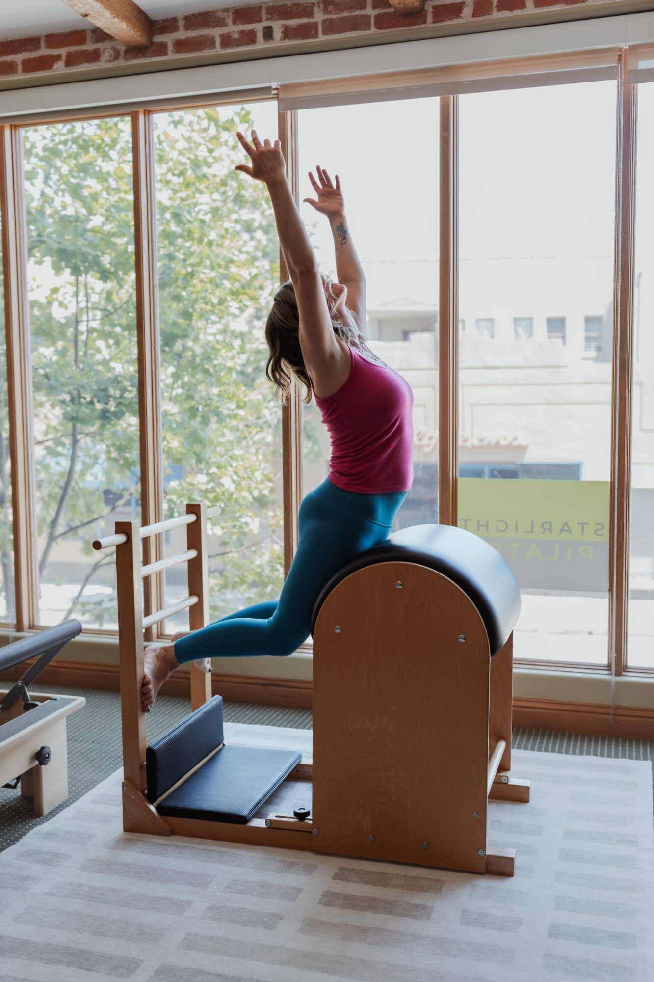 Person in a pink top and blue pants practicing Pilates on a reformer machine in a bright room with large windows.