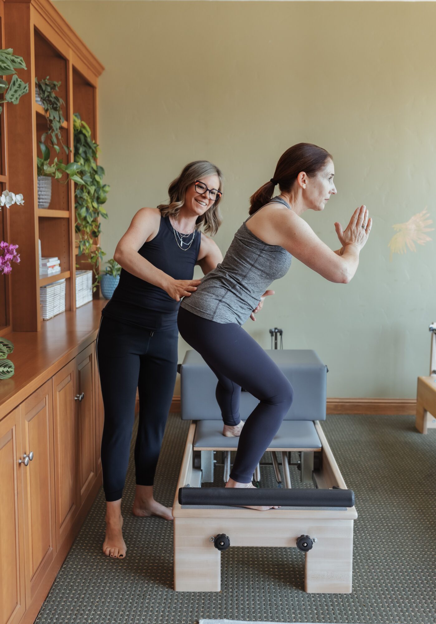Two women in a room, one standing and the other kneeling on a small wooden platform, engaging in an activity. A wooden cabinet with plants is on the left, and a green wall is in the background.