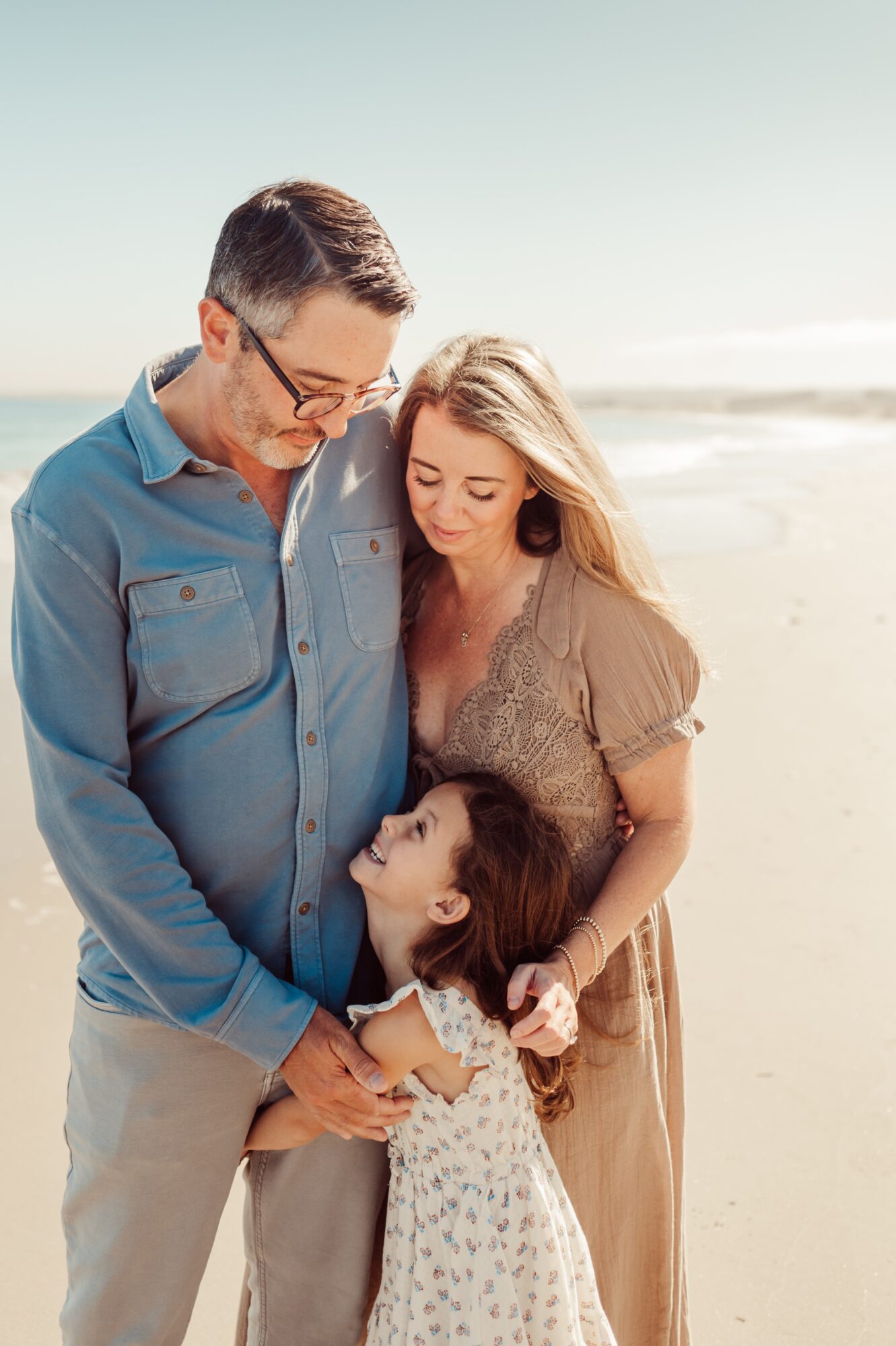 Family of three on the beach, father, mother, and daughter, smiling and holding hands, with ocean in background.